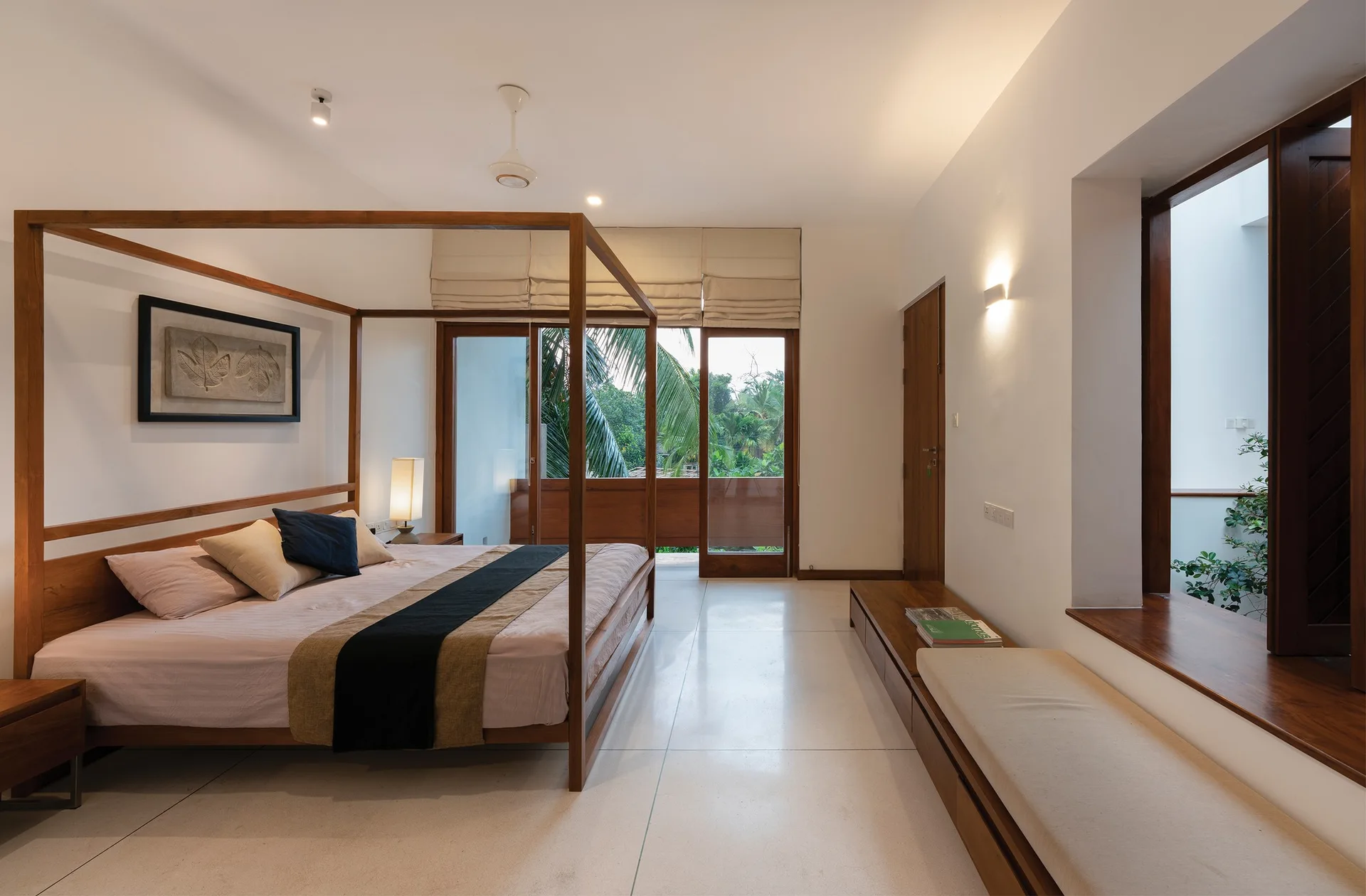 A bright minimalist bedroom features a wooden four-poster bed, a long built-in wooden bench, and large glass doors opening to lush green trees.