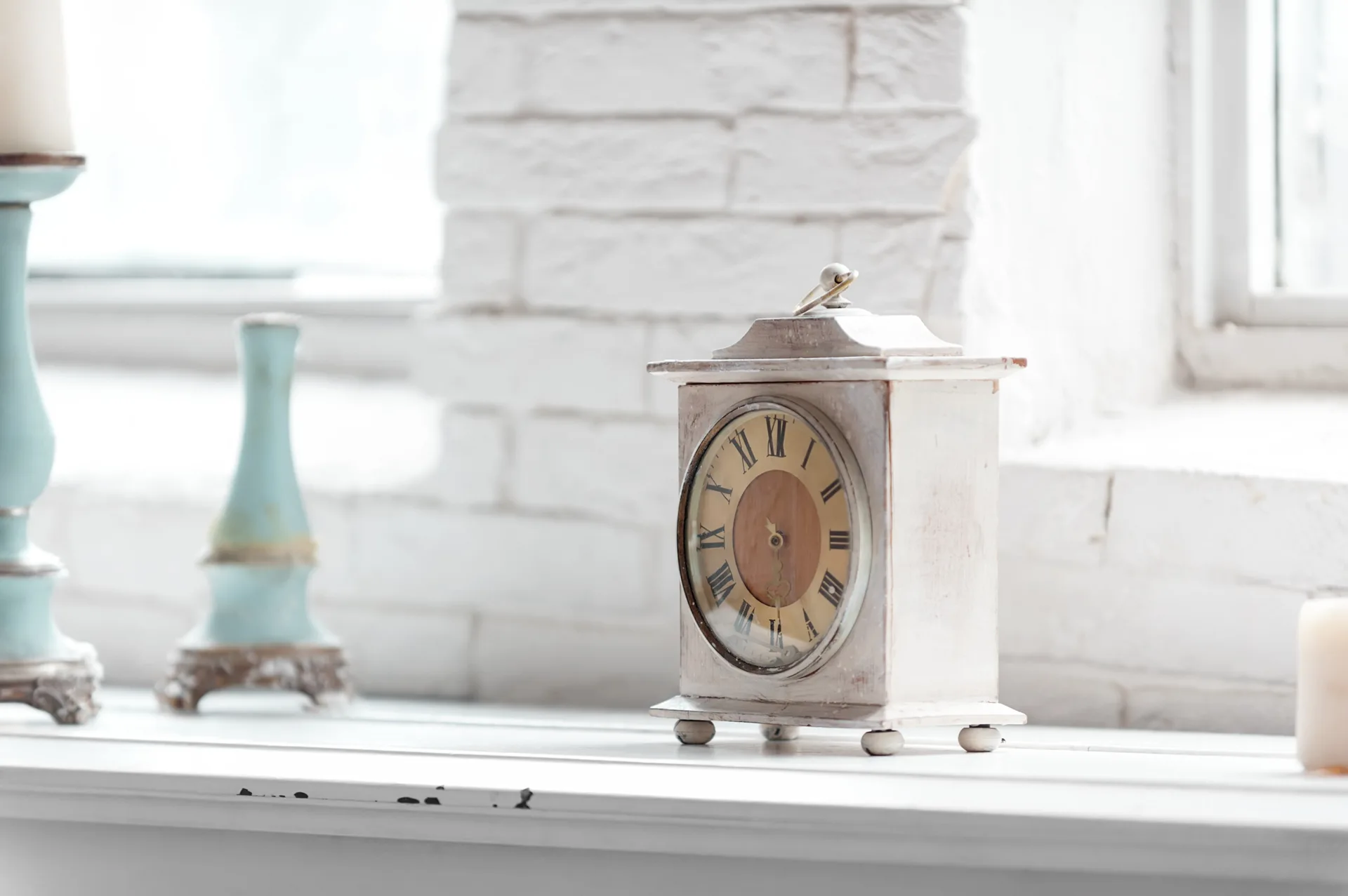 A vintage white table clock with Roman numerals sits on a white windowsill next to light blue candlesticks in front of a painted brick wall.