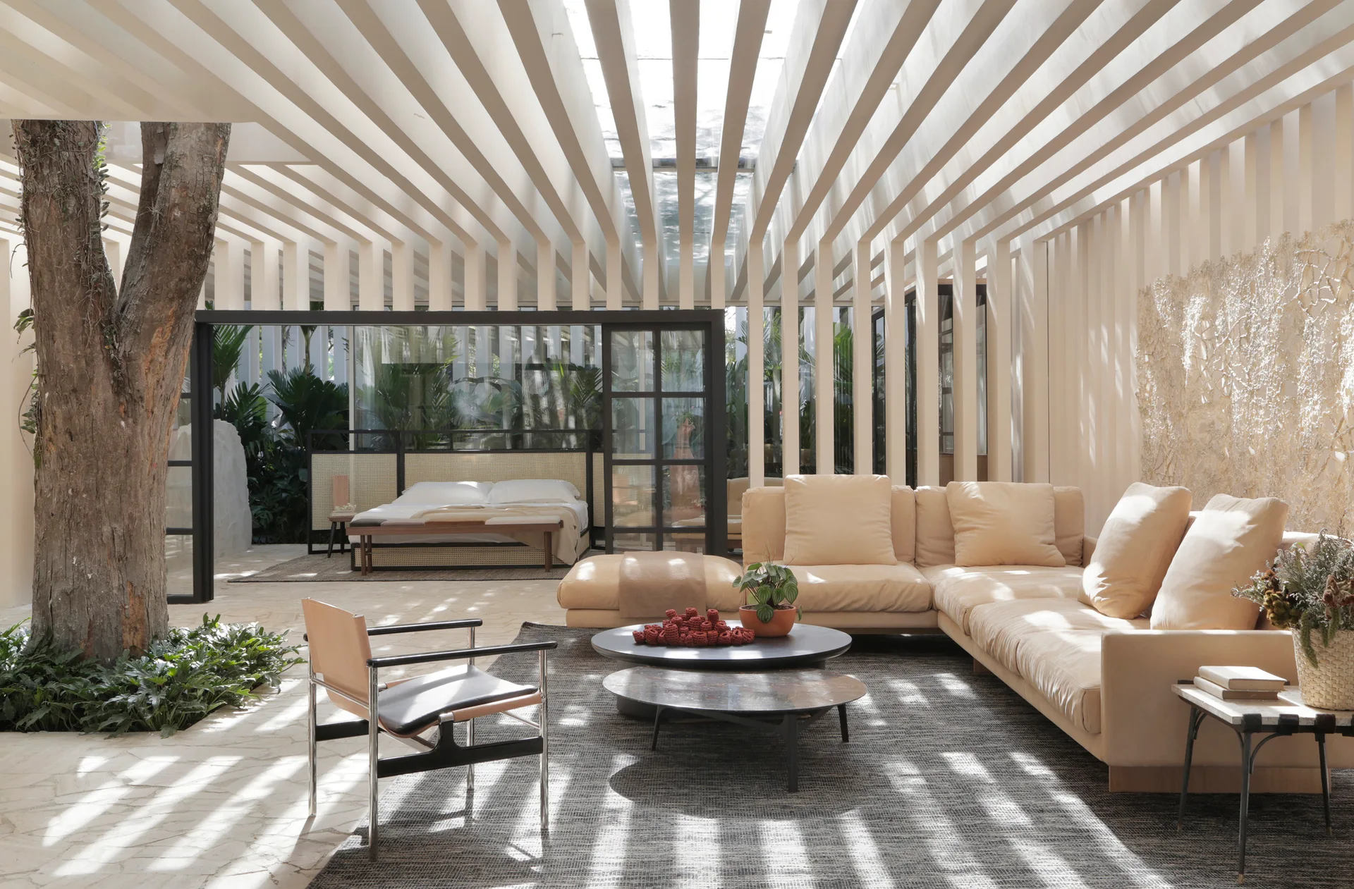 A sunlit living room features a cream sectional sofa, a large indoor tree, and rhythmic ceiling beams that cast linear shadows on the stone floor.