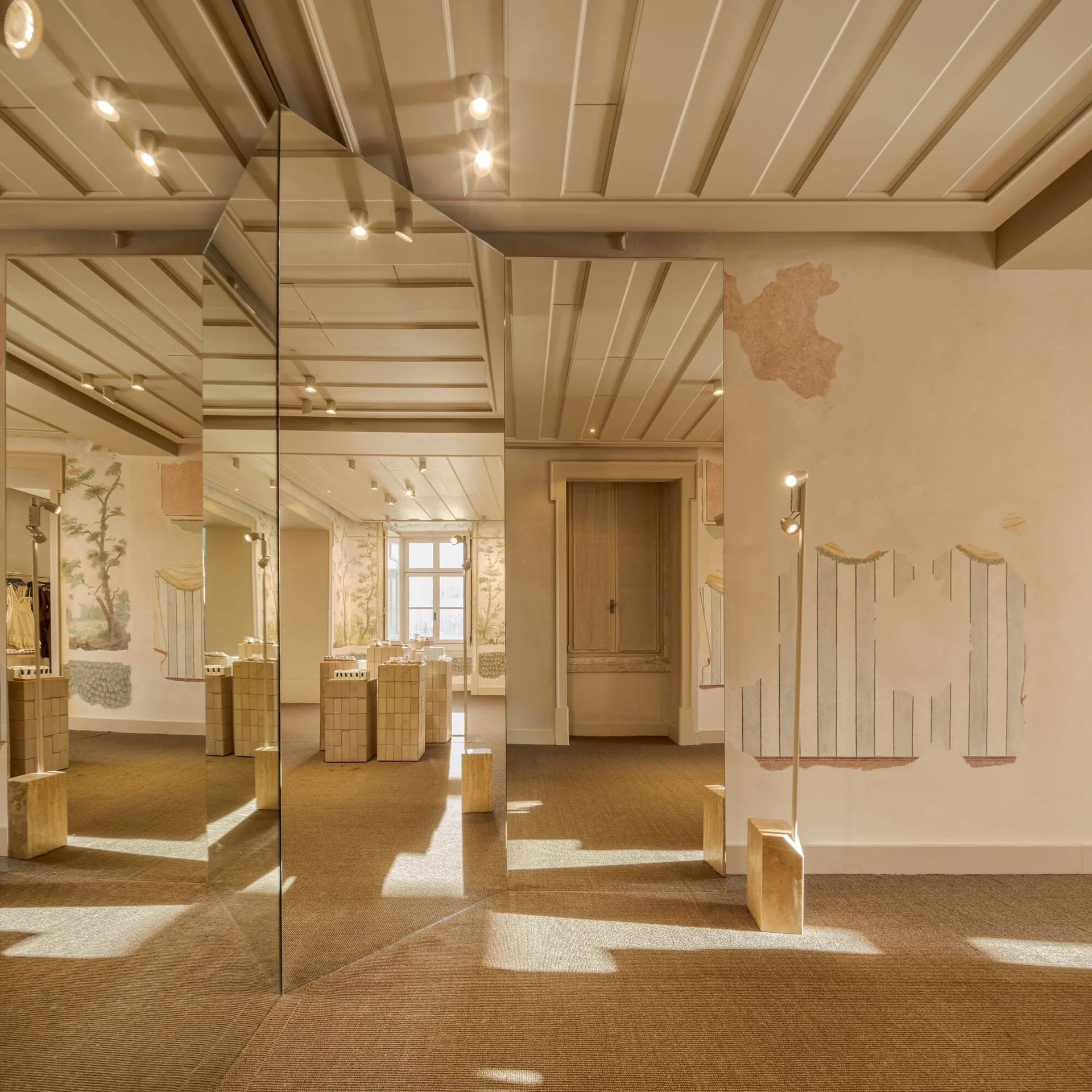 A minimalist room features large angled mirrors reflecting modular wooden blocks alongside distressed walls with faded frescoes.