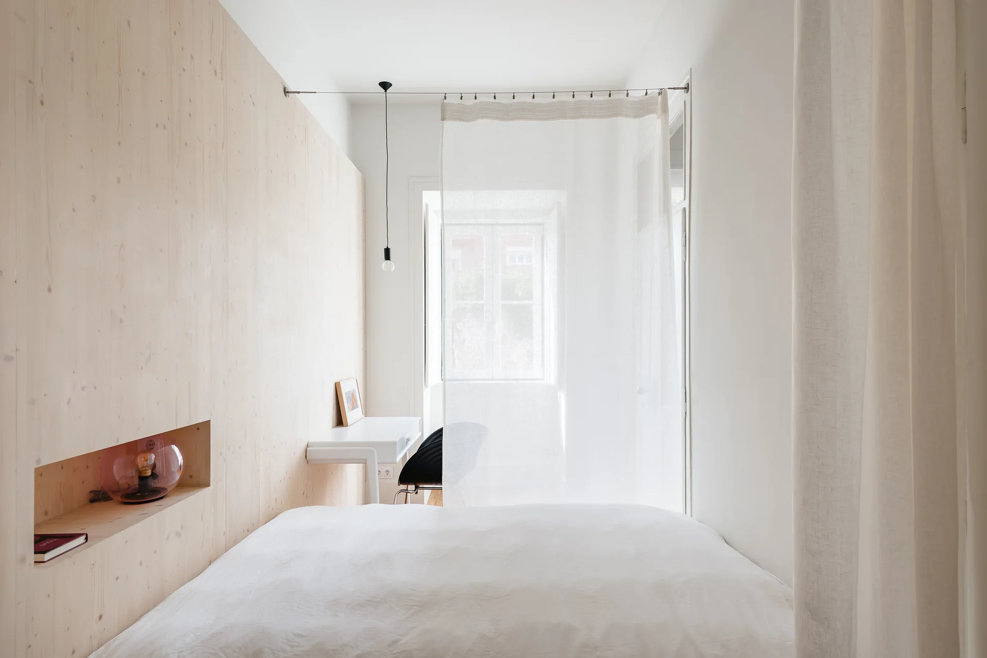 A minimalist bedroom features a light wood-paneled wall with a built-in alcove, white bedding, and a sheer curtain room divider near a desk.
