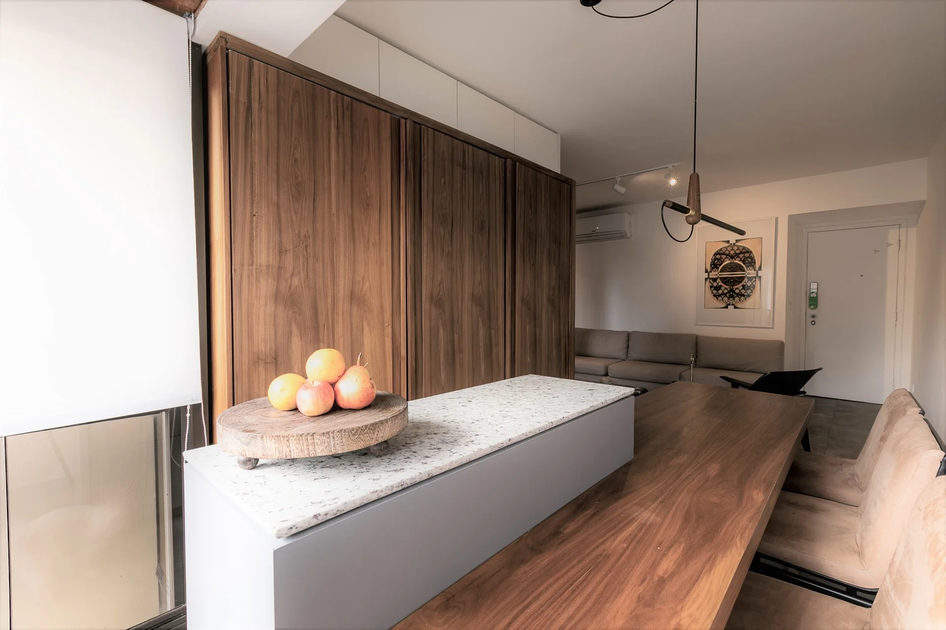A modern dining space features a long wood table paired with a gray stone-topped island next to floor-to-ceiling wood cabinets.