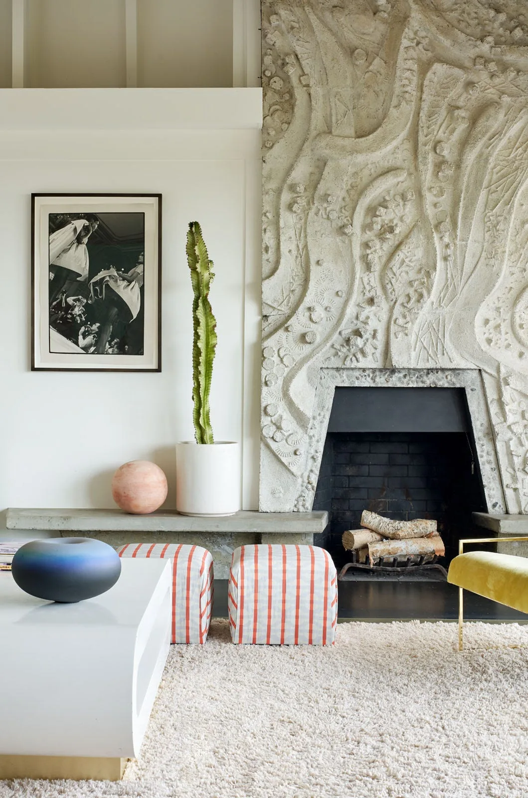 A modern living room features a highly textured sculptural stone fireplace surround, striped ottomans, a shaggy cream rug, and a tall potted cactus.