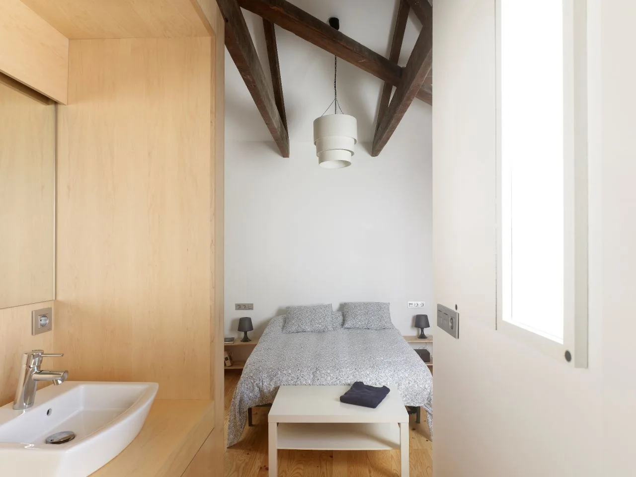 A minimalist bedroom featuring exposed dark wood ceiling beams, a bed with patterned grey bedding, and an integrated light wood vanity with a white sink.