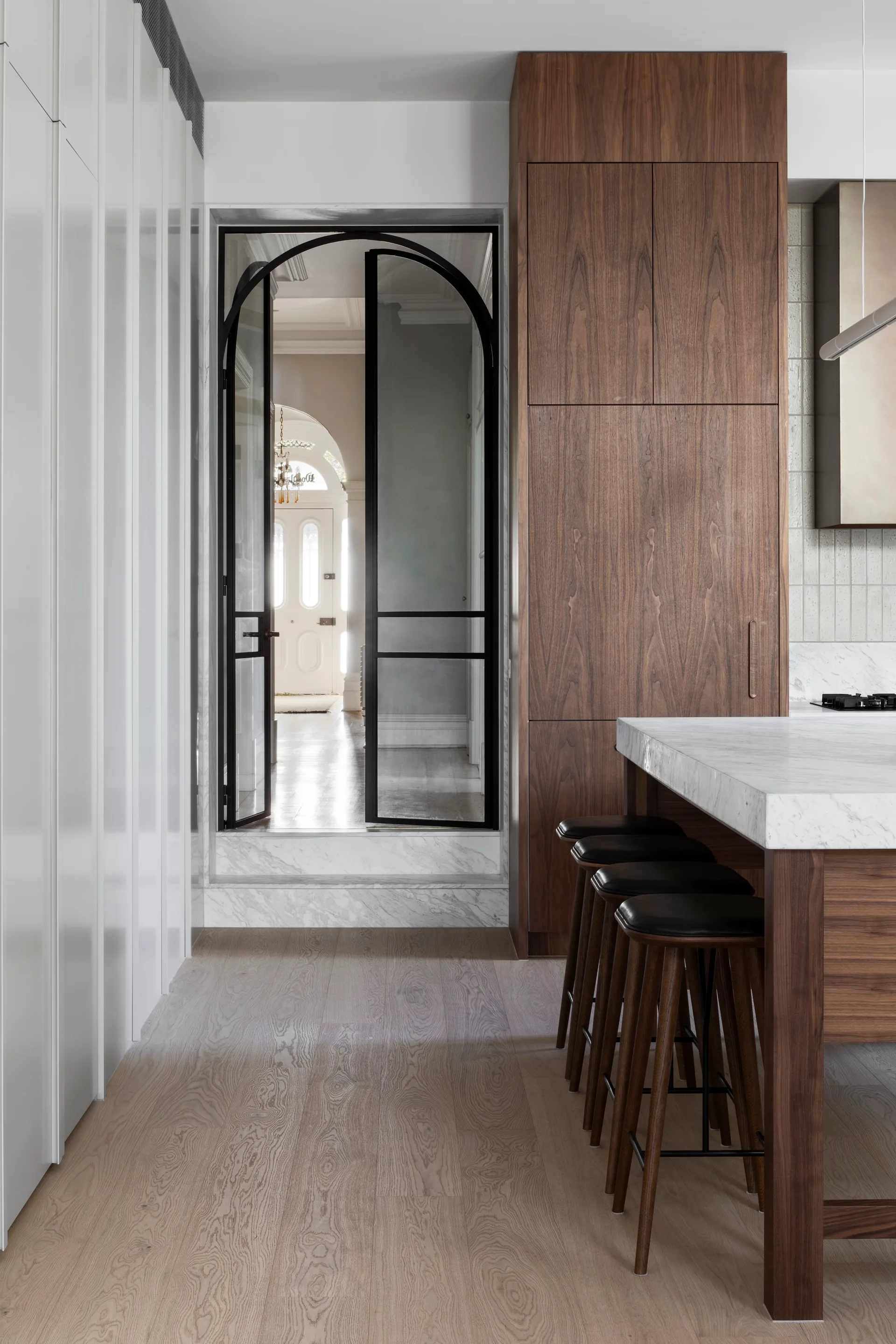 A modern kitchen featuring a walnut wood island with a white marble countertop, dark bar stools, and an arched black-framed glass door leading to a hallway.