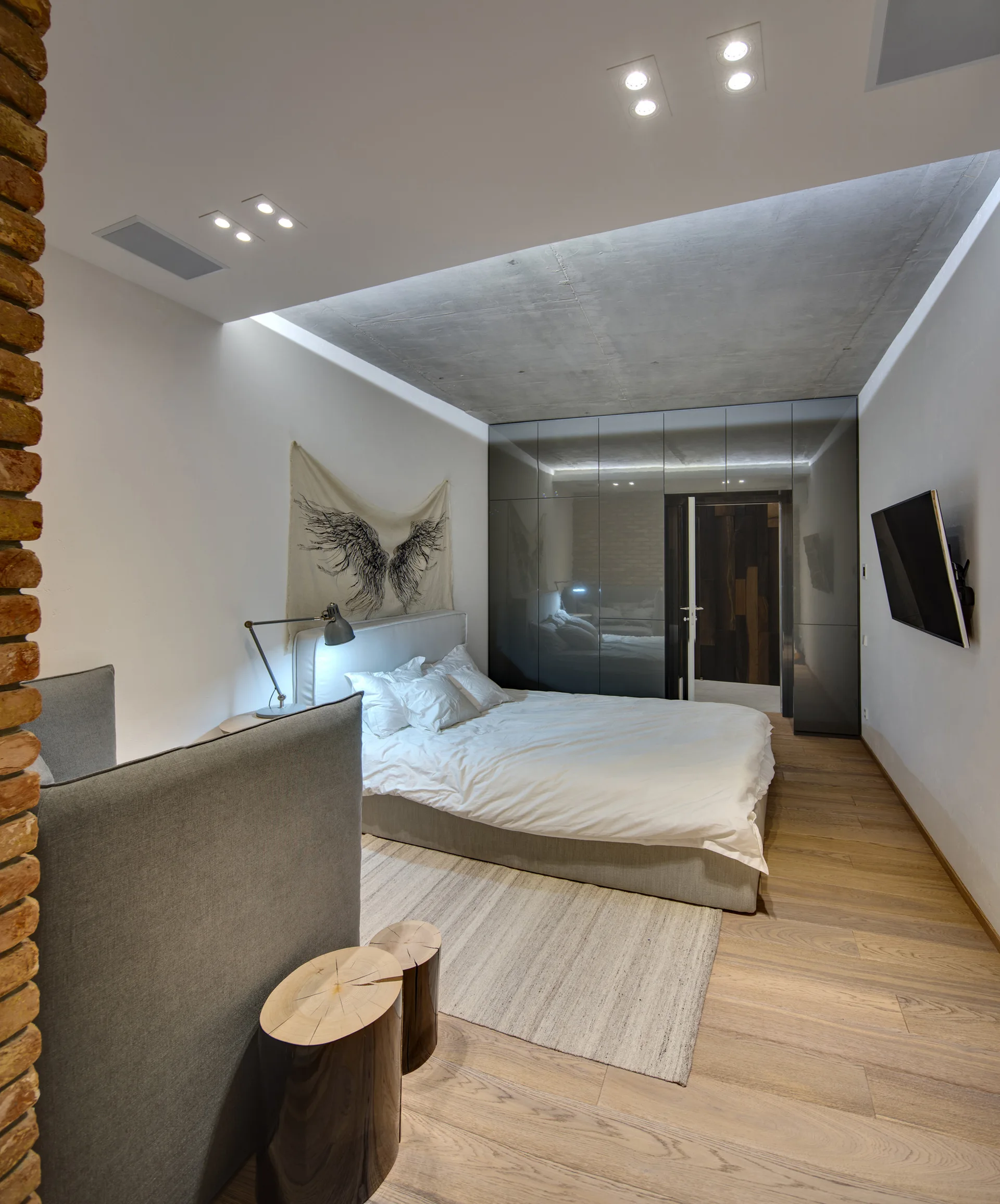 A modern bedroom featuring a low platform bed with white sheets, a large glossy grey built-in wardrobe, and a raw concrete ceiling insert.