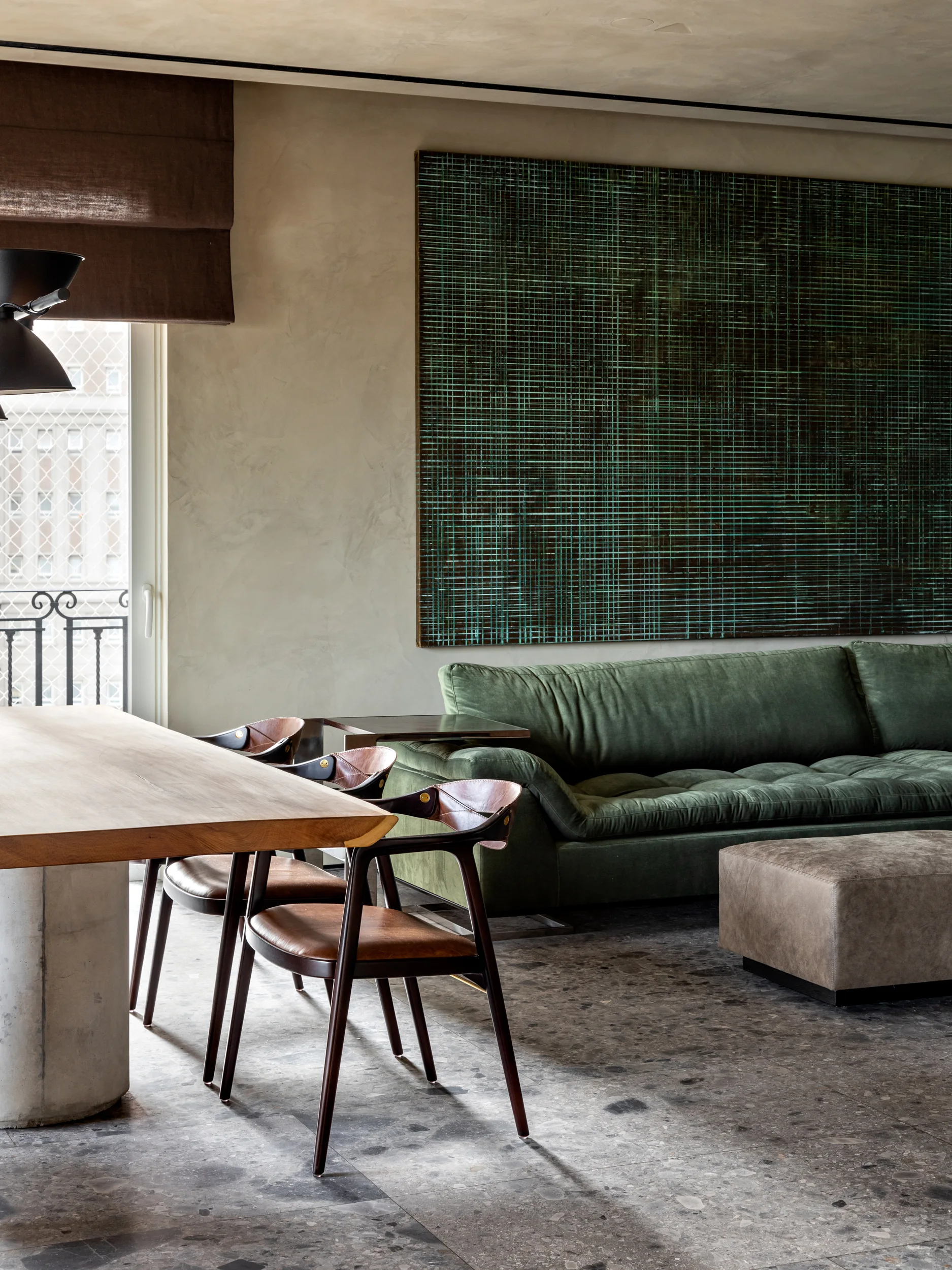 A contemporary living room features a green tufted sofa beneath a large abstract green painting, alongside a wooden dining table with leather chairs.