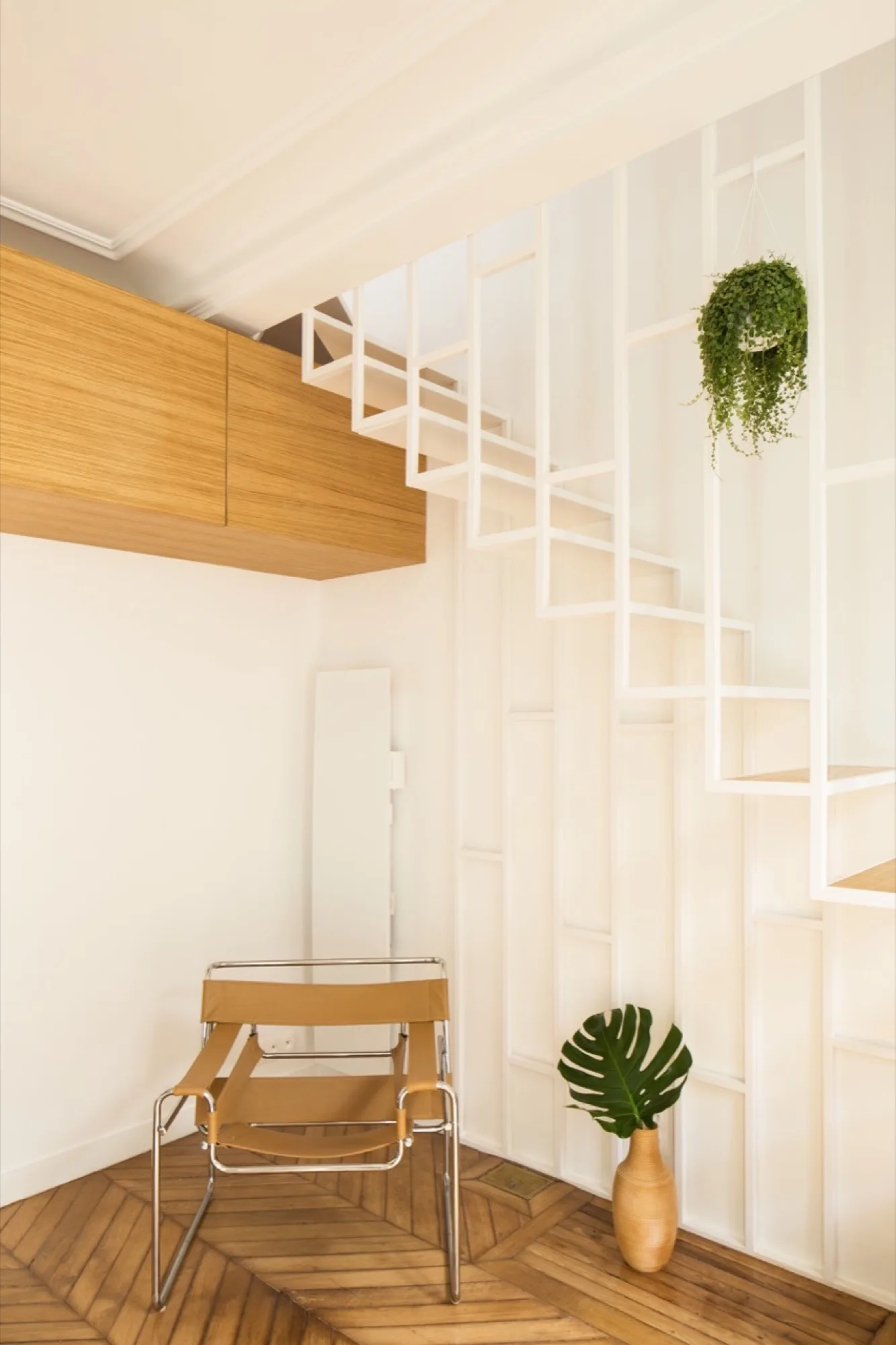 An open white geometric staircase rises above a tan leather sling chair on herringbone pattern wood flooring.