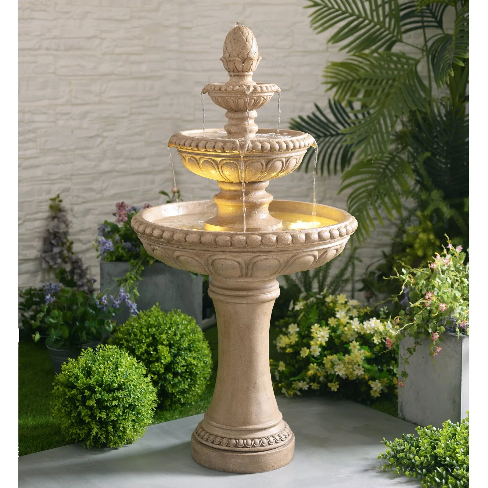 A three-tiered tan stone-like water fountain with glowing warm lights stands on a grey patio surrounded by potted green plants and a textured wall.