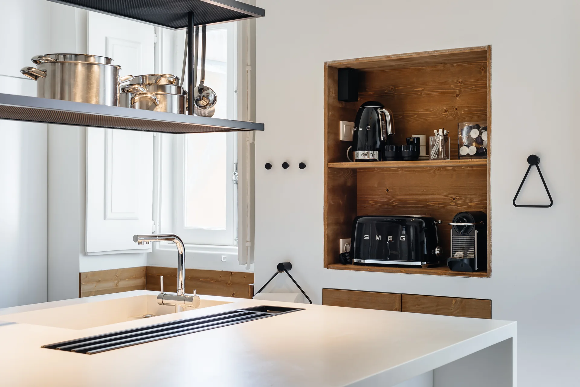 A modern white kitchen island with a chrome faucet stands in front of a white wall with a recessed wooden niche housing black small appliances.