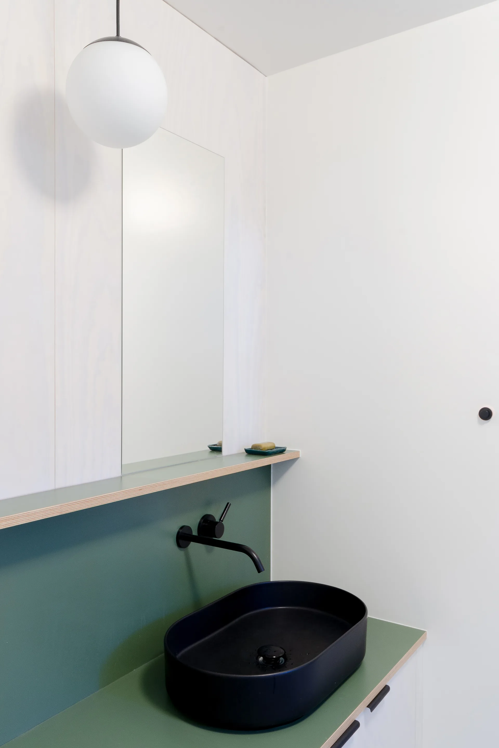 A minimalist bathroom features a black oval vessel sink on a sage green countertop, a wall-mounted black faucet, and a frameless mirror.