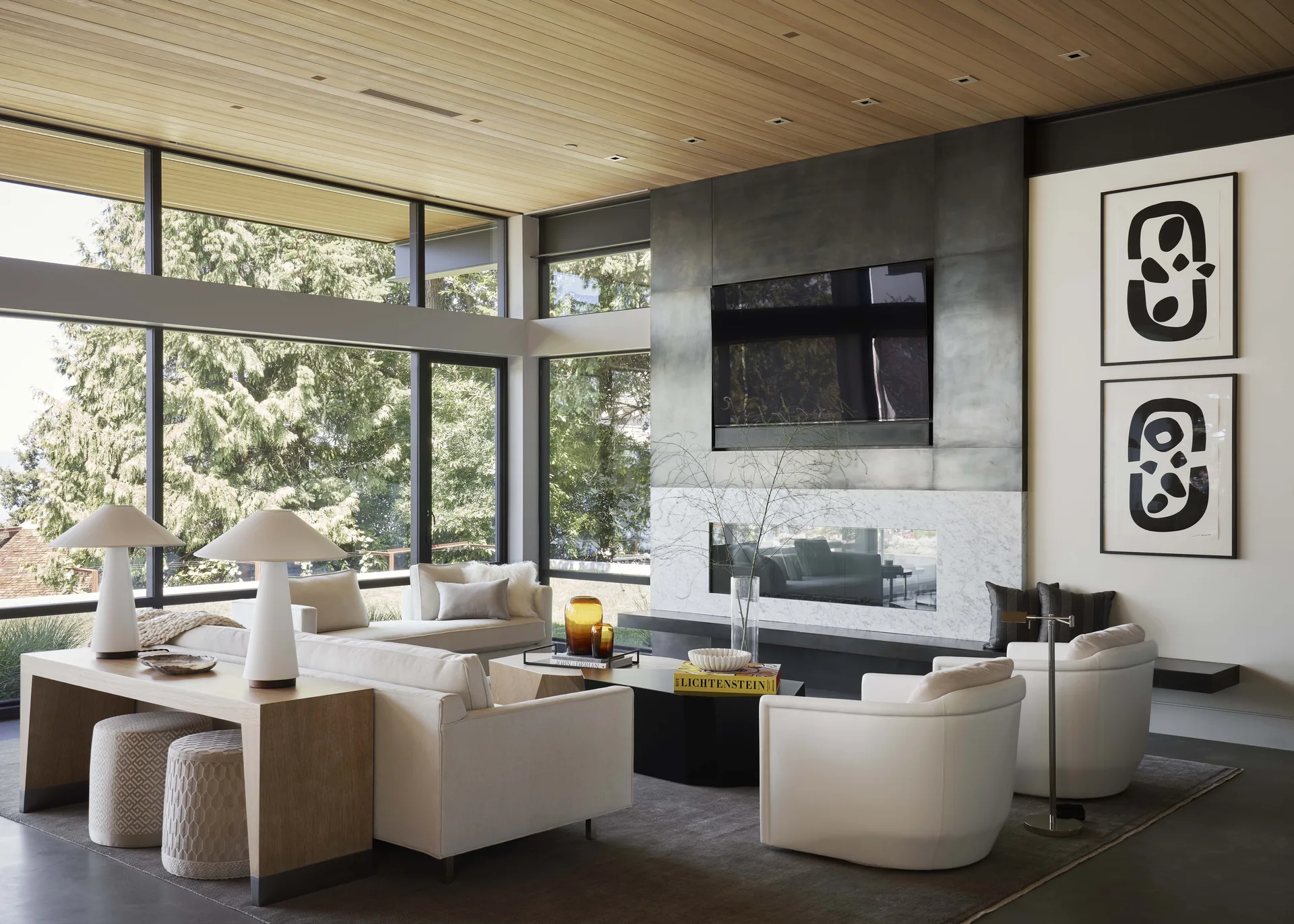 Modern living room featuring a wood-planked ceiling, a dark metal fireplace surround, white sofas, and large windows displaying forest views.
