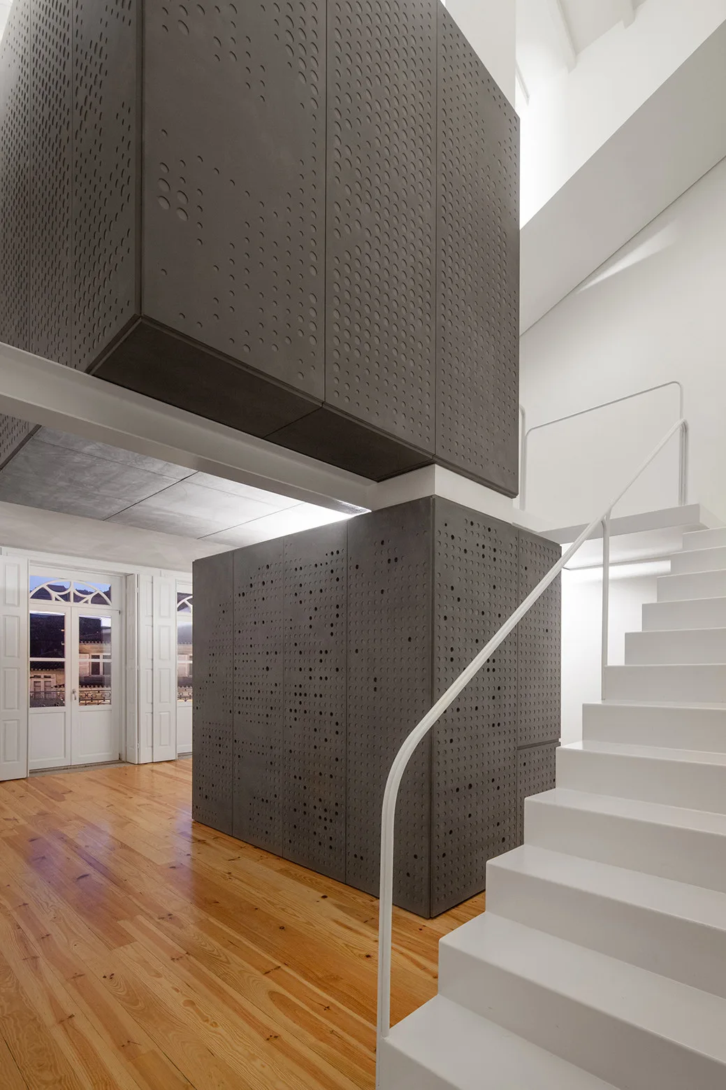 A minimalist interior featuring a white staircase with a metal railing alongside a central charcoal gray storage unit made of perforated panels.