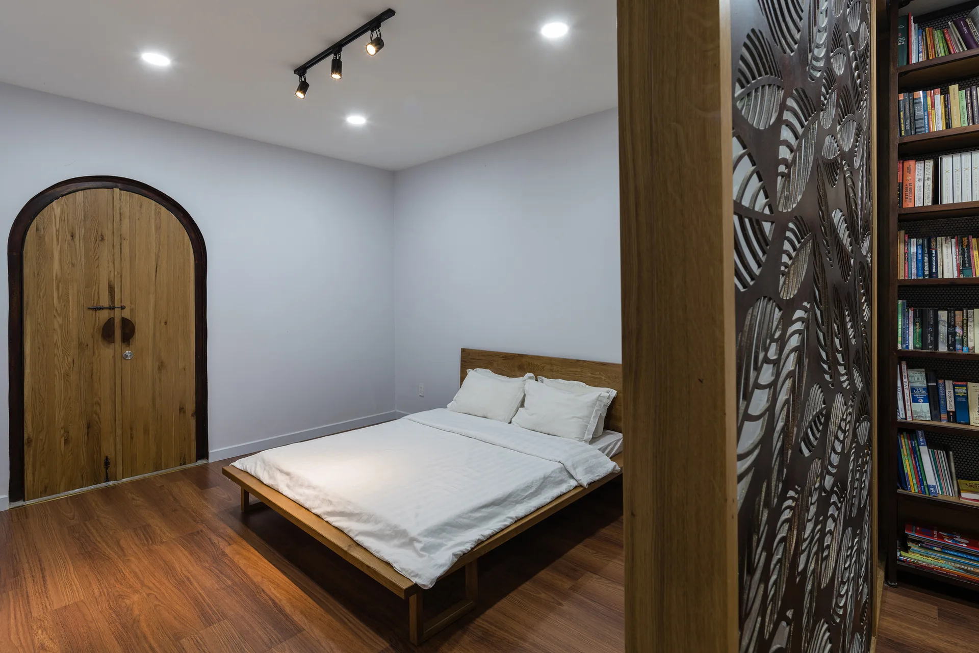 A minimalist bedroom featuring a low wood platform bed, an arched wooden door, and a decorative leaf-patterned wood screen in the foreground.