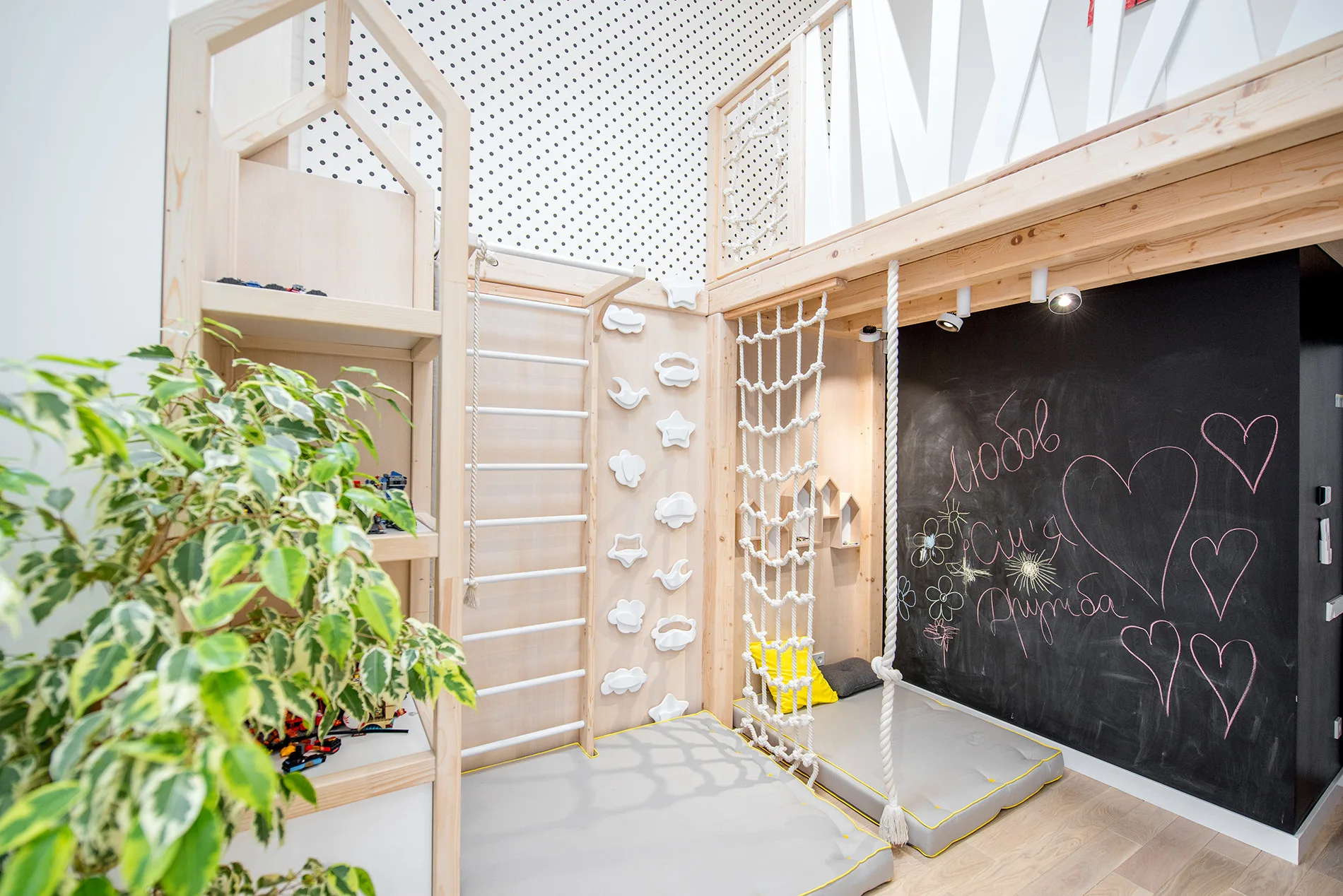 An indoor kids play area features a light wood climbing loft, a climbing wall, grey floor mats, and a large black chalkboard wall.