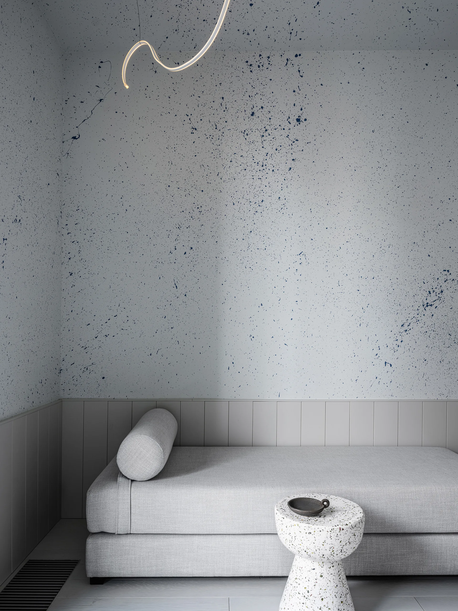 A grey upholstered daybed sits against a white wall featuring a blue paint splatter pattern, accompanied by a small terrazzo side table.