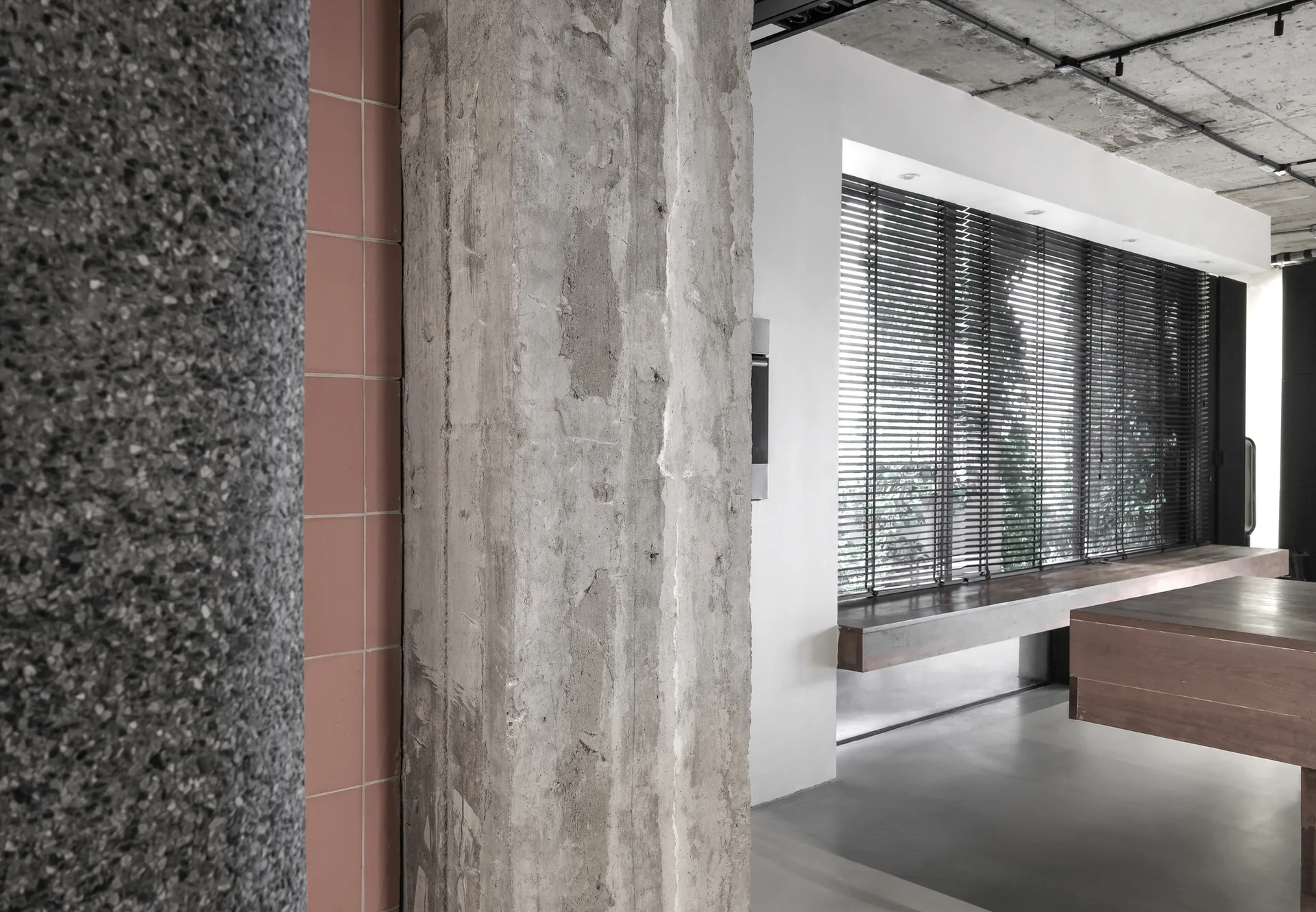 An industrial interior room features a prominent raw concrete column, a dusty pink tiled accent wall, and a large window with dark horizontal blinds.
