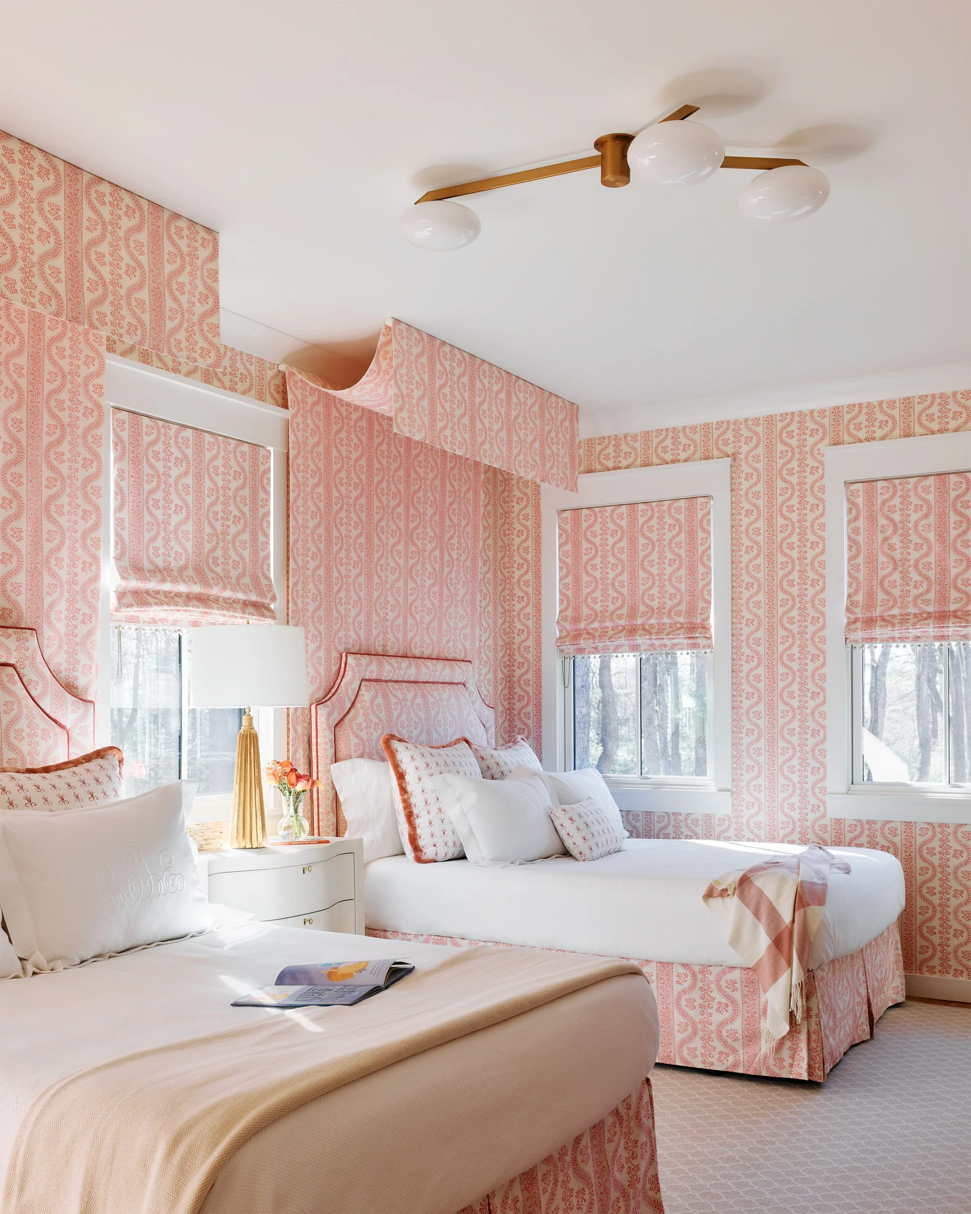 Two twin beds feature matching pink and white patterned headboards, coordinating with the wallpaper and roman shades in a bright guest bedroom.