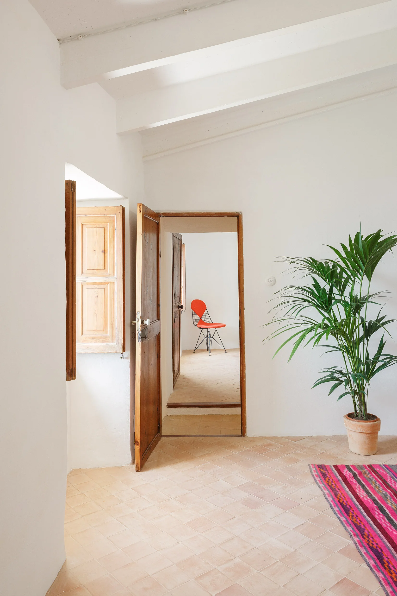 A bright white hallway with terracotta tiled flooring, rustic wooden doors, an indoor palm plant, and a distant orange chair seen through a doorway.