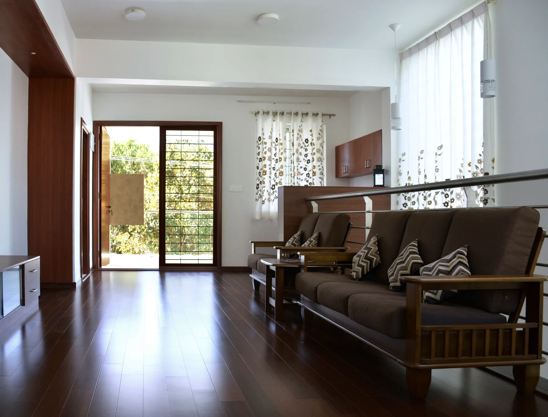 A spacious living room features polished dark wood flooring, brown wooden-framed sofas, a metal railing, and a large sliding glass door.