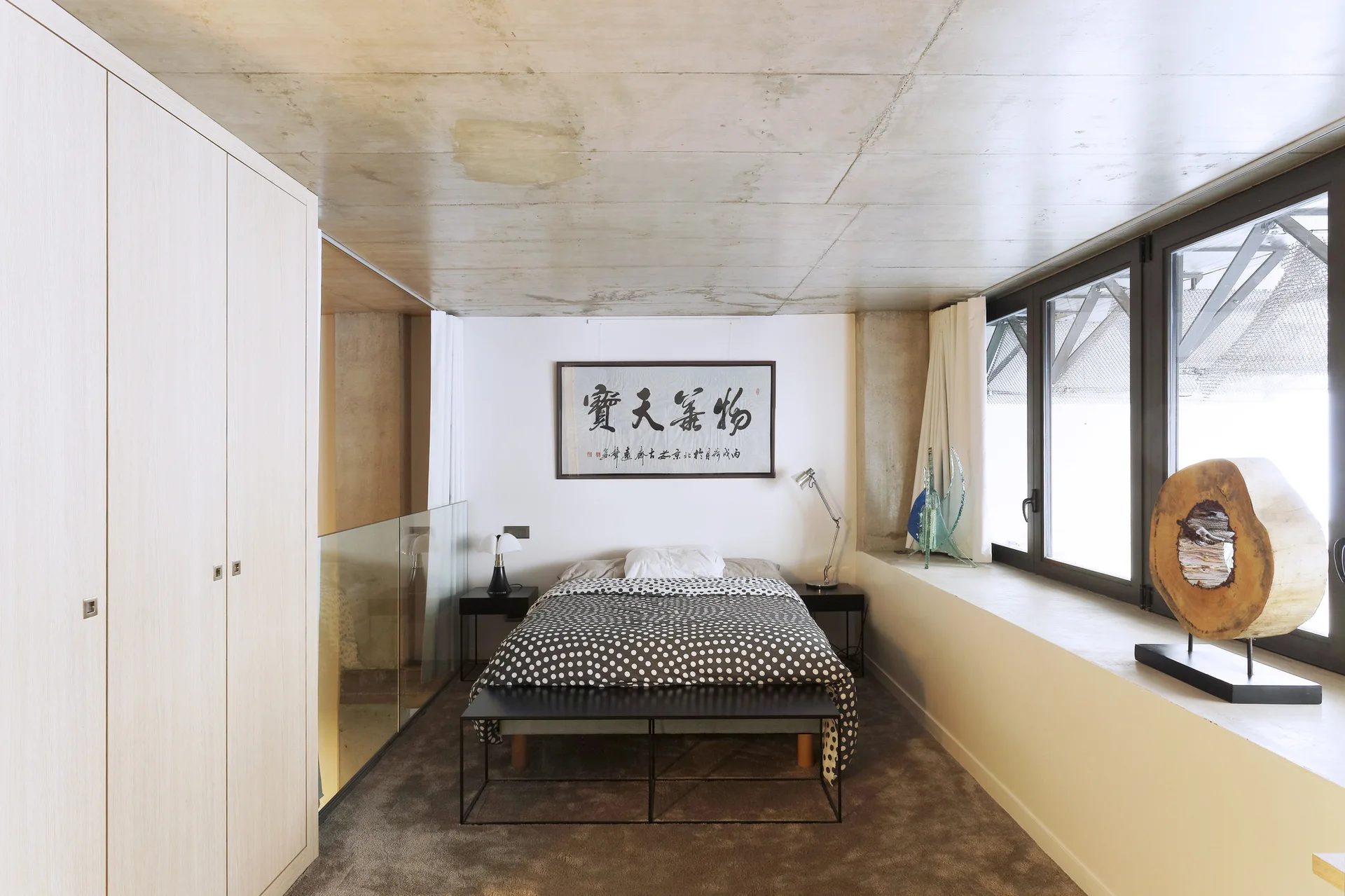 A modern loft bedroom with polka dot bedding, a concrete ceiling, a light wood wardrobe, and a wide window ledge.