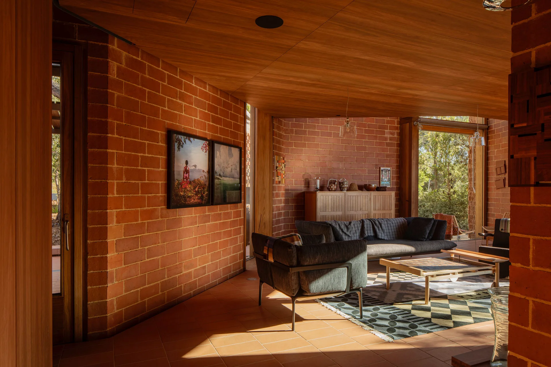 A living room features exposed red brick walls, a wood paneled ceiling, a dark sofa, and geometric patterned rug on terracotta tiles.