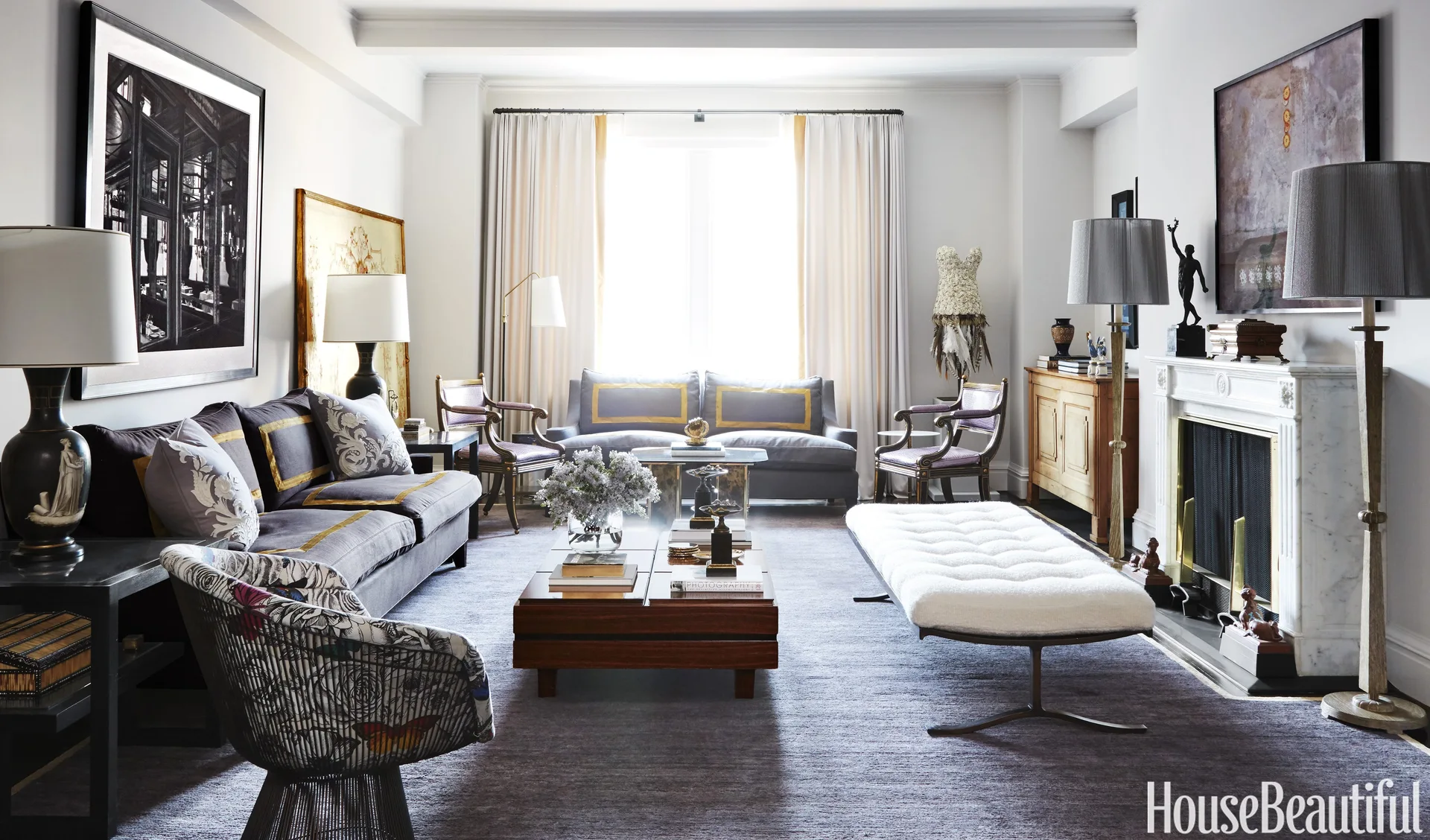 An eclectic living room featuring gray sofas with gold trim, a white tufted bench, a wooden coffee table, and a white marble fireplace.