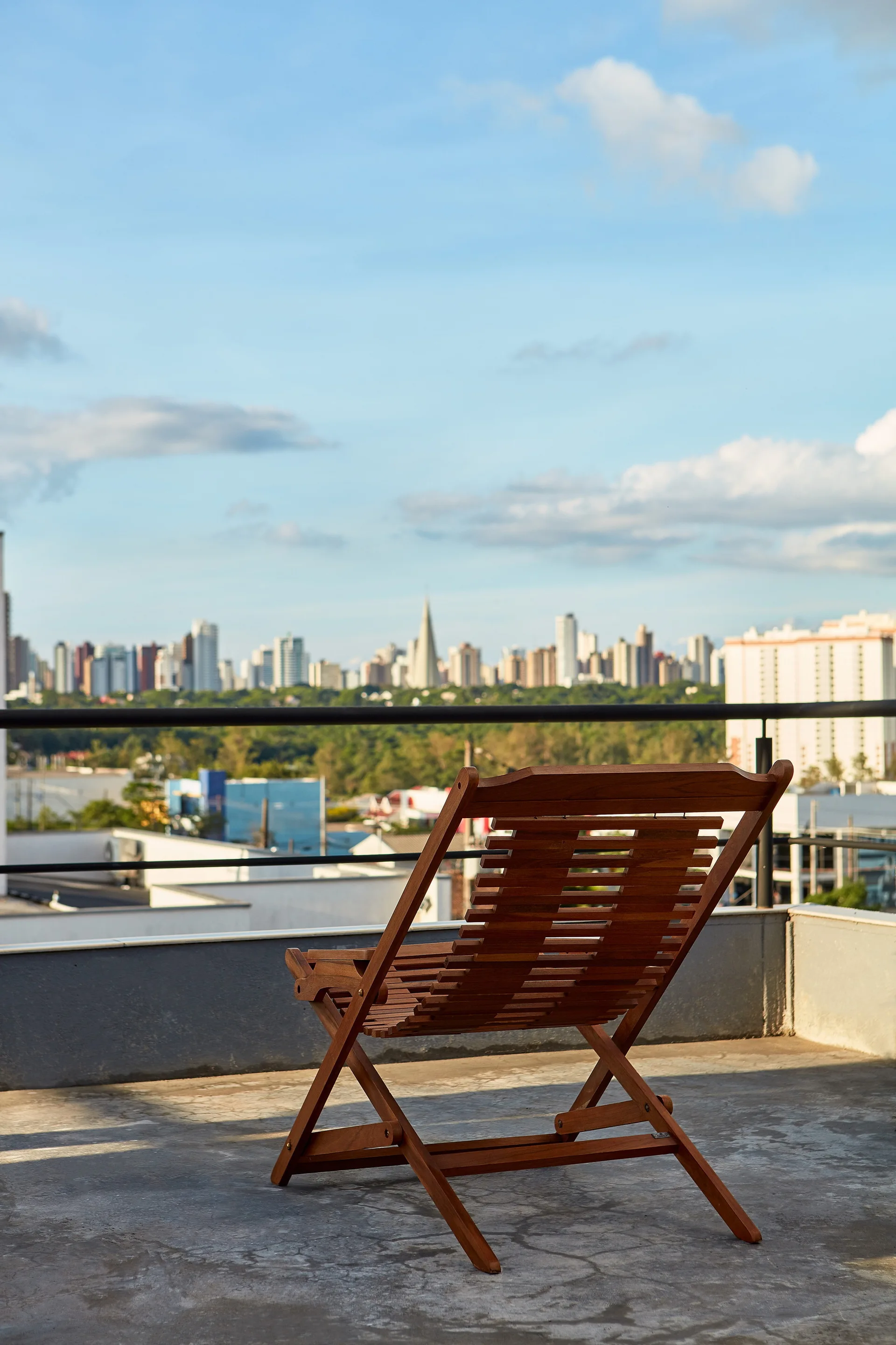A slatted wooden deck chair sits on a grey concrete balcony with a black metal railing overlooking a cityscape under a blue sky.