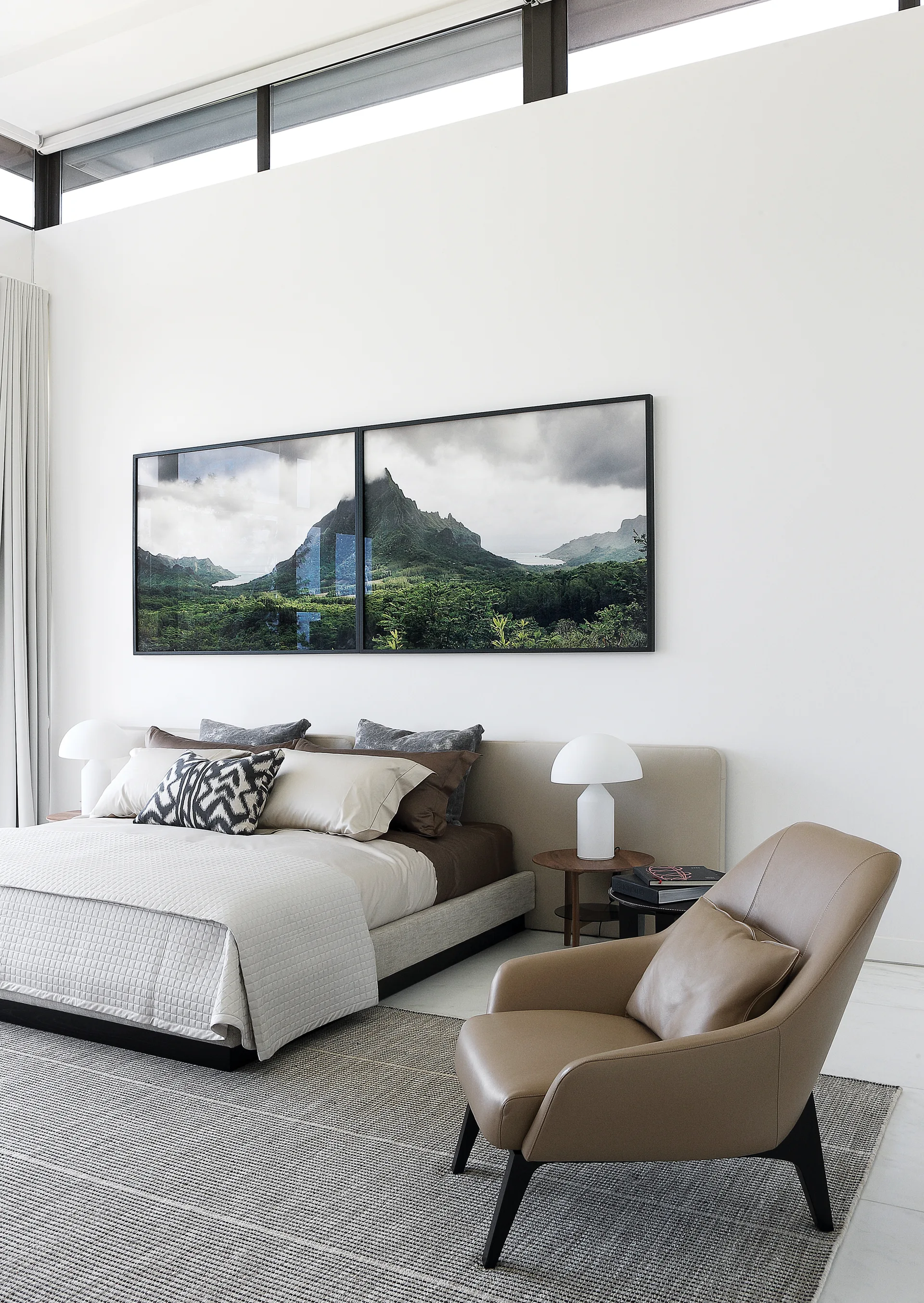 A modern bedroom featuring a low platform bed with earth-toned bedding, a tan leather armchair, and large nature-themed wall art below high windows.