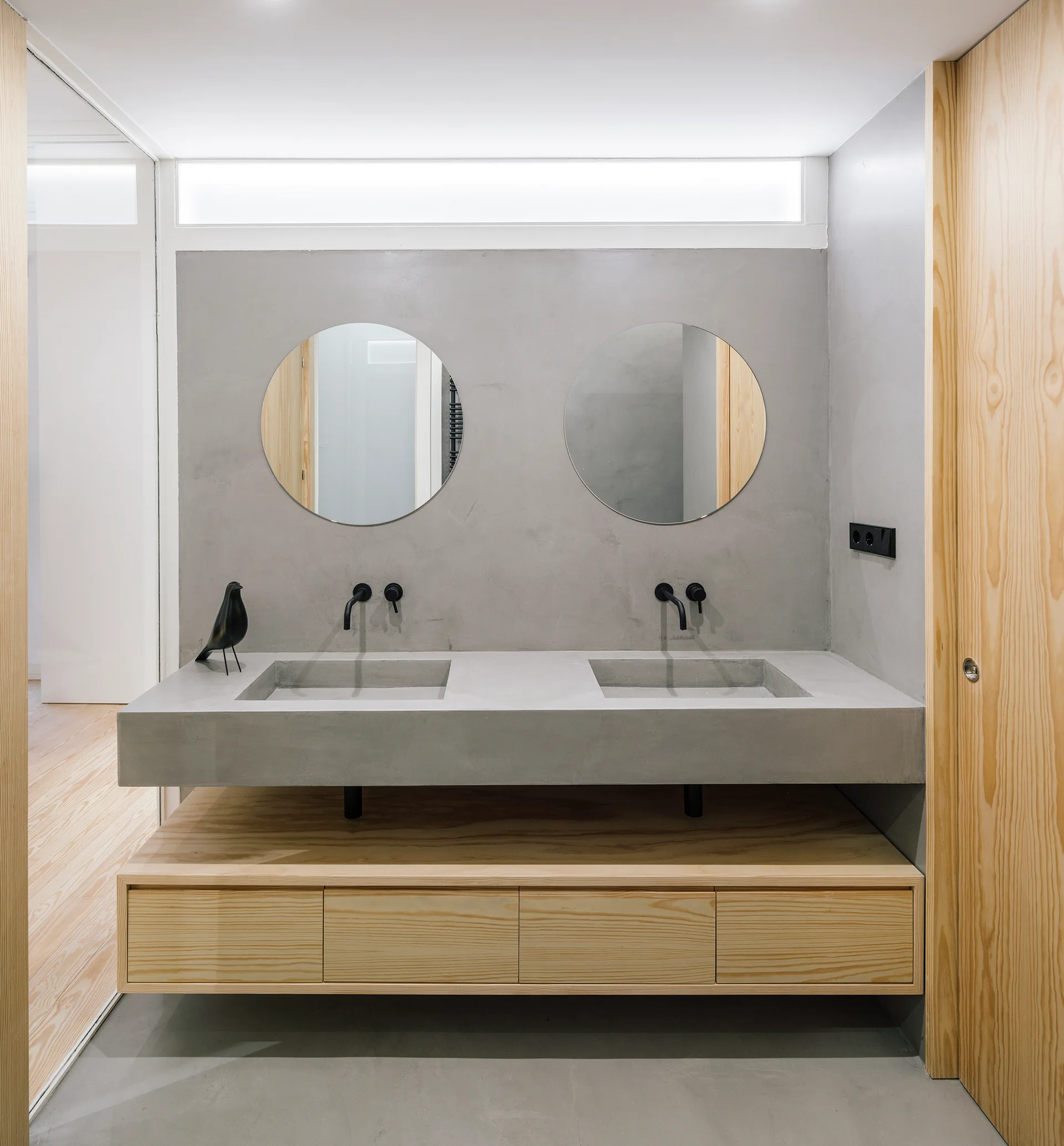 A minimalist bathroom features a floating double concrete vanity, two circular mirrors, matte black wall-mounted faucets, and a light-wood lower cabinet.