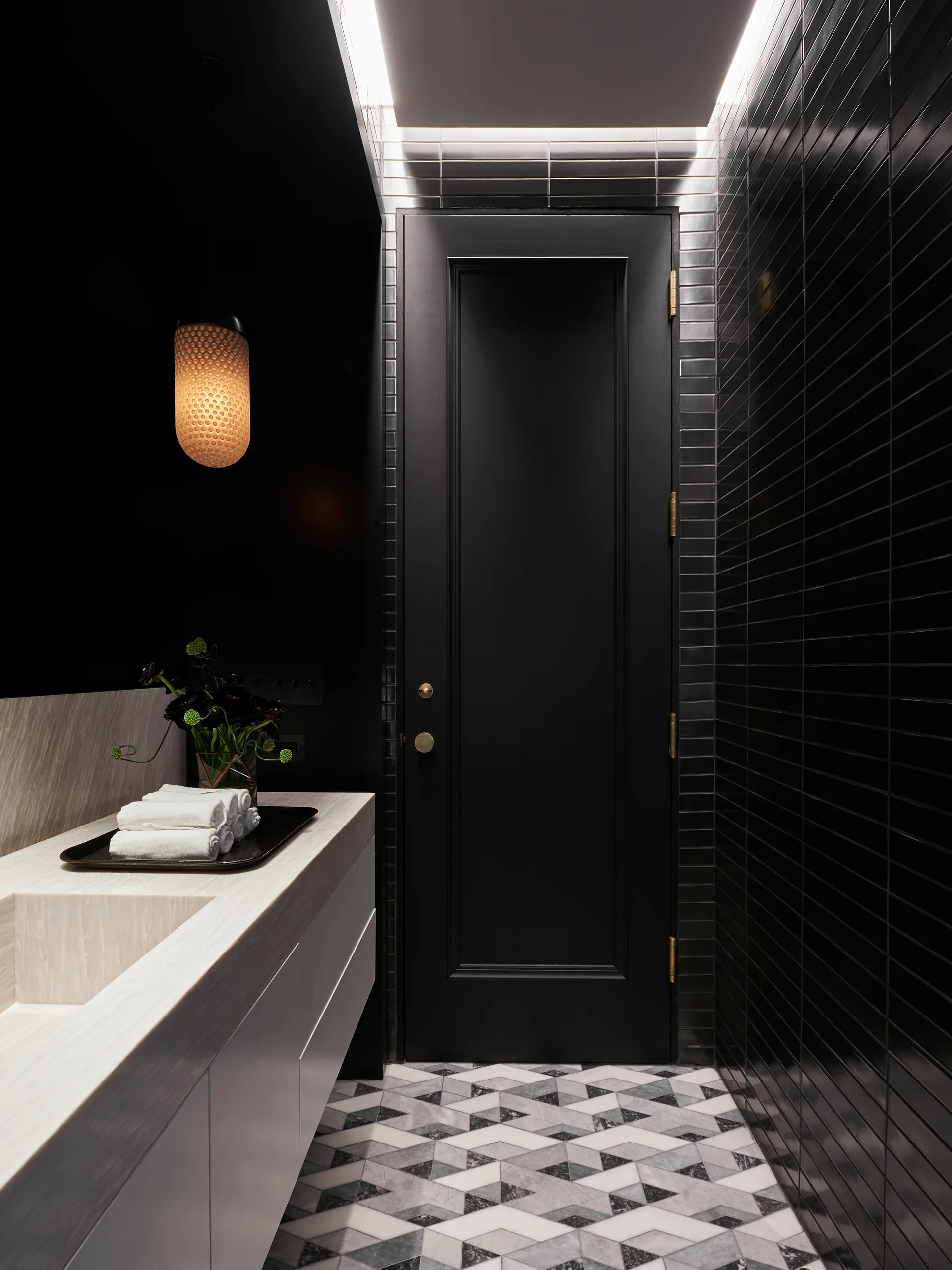 A modern powder room featuring a light stone vanity, glossy black vertical subway tile walls, a matte black door, and geometric floor tiles.