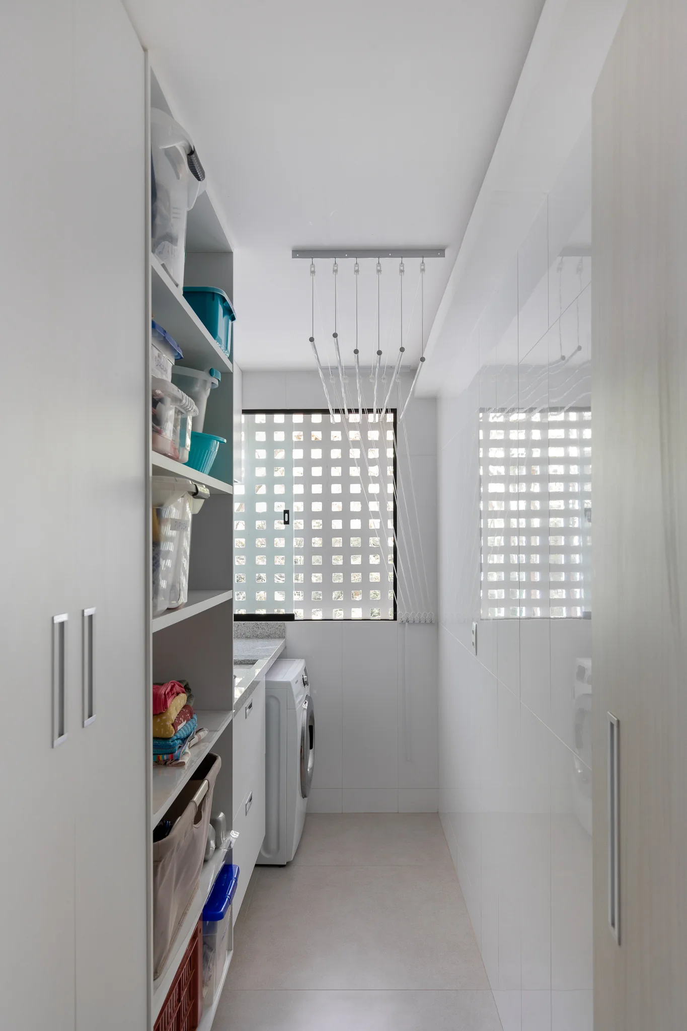A narrow white laundry room features tall built-in shelving, a front-loading washing machine, and an overhead ceiling-mounted drying rack.
