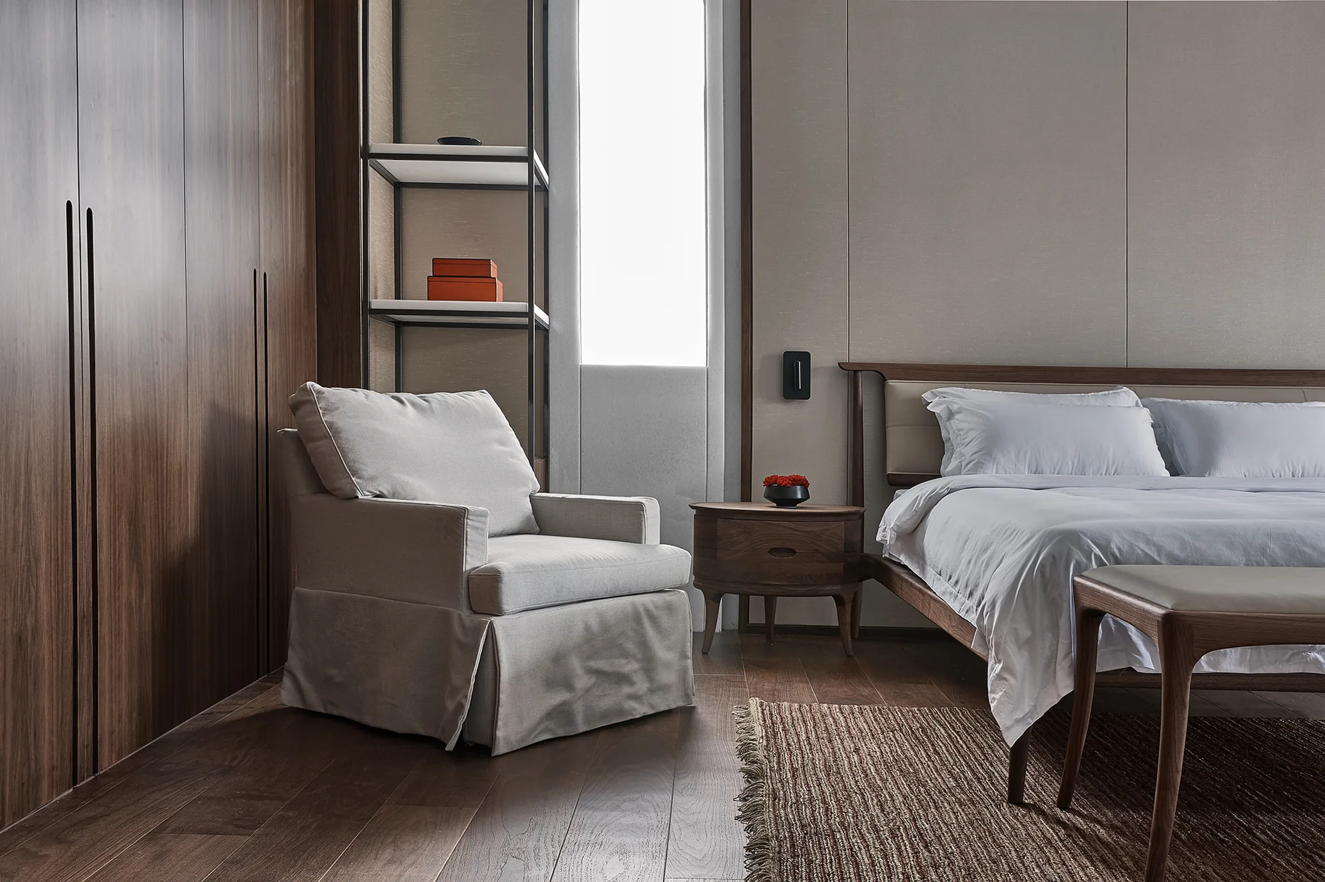 A contemporary bedroom featuring a dark wood wardrobe, a light gray upholstered armchair, a wood bed with white linens, and a tall metal shelving unit.