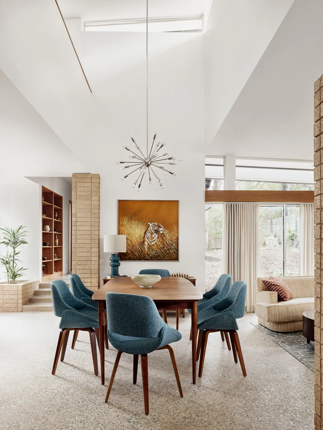 A mid-century dining space features a wooden table with blue chairs, a sputnik chandelier, and brick pillars on a speckled terrazzo floor.