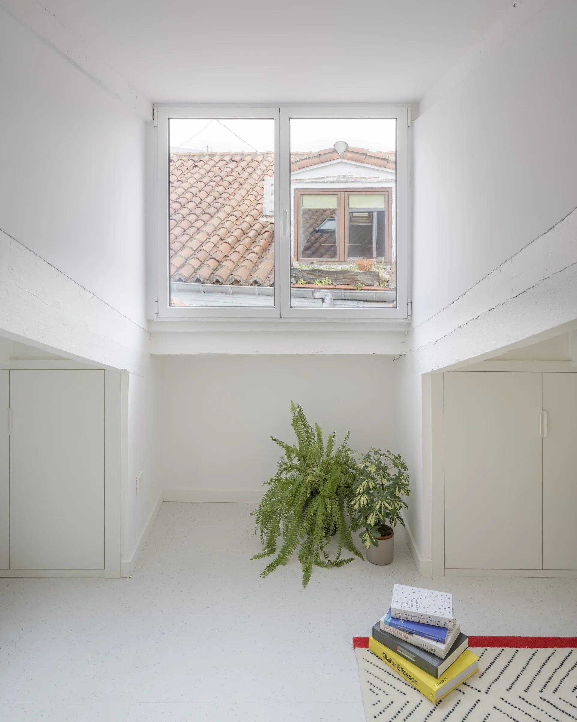 A bright white attic alcove featuring symmetrical built-in cabinets, a central window looking out at terracotta rooftops, and potted plants on a speckled floor.