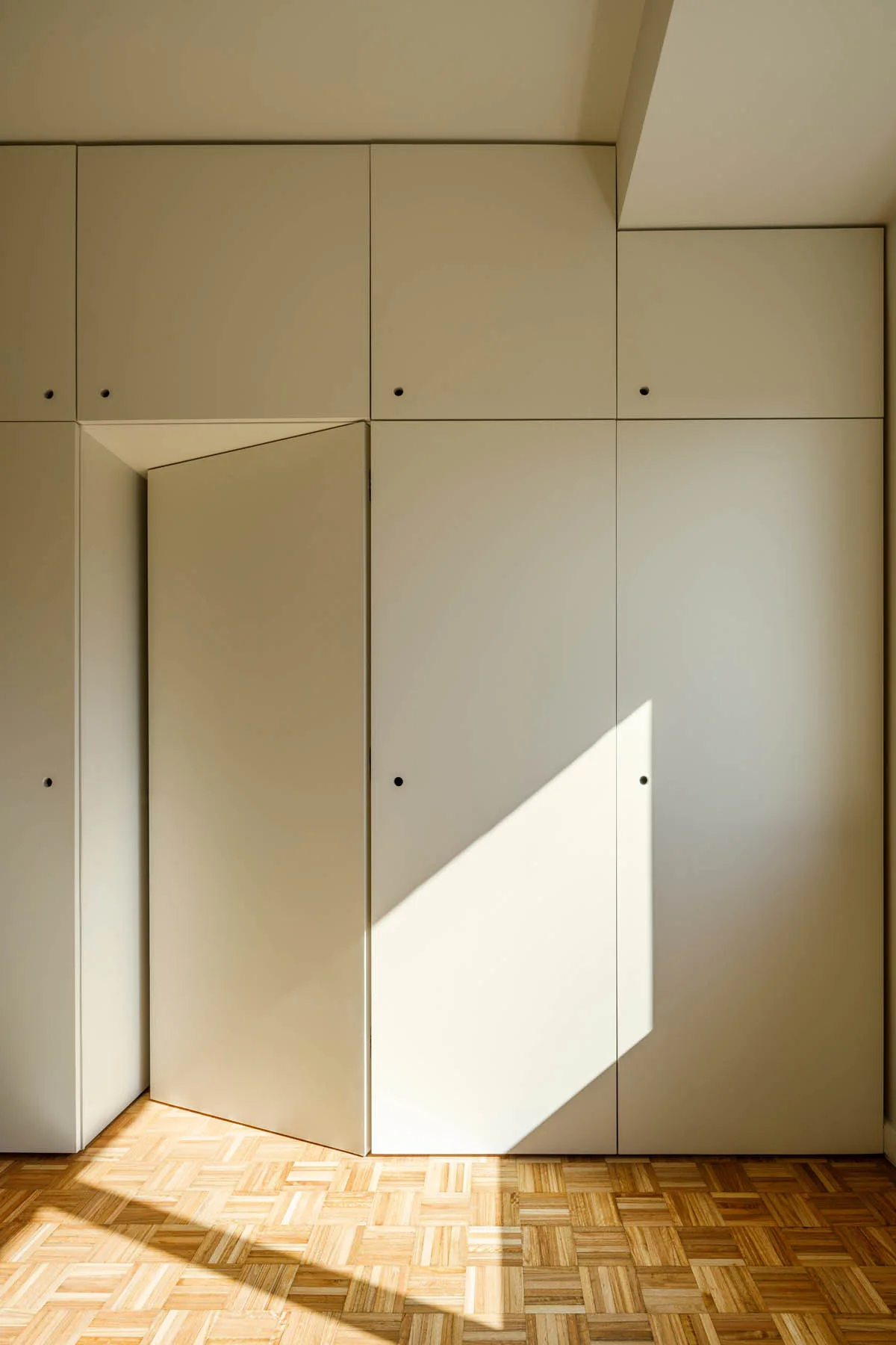 Cream floor-to-ceiling minimalist storage cabinets with small round finger pulls sit above a geometric parquet wood floor.