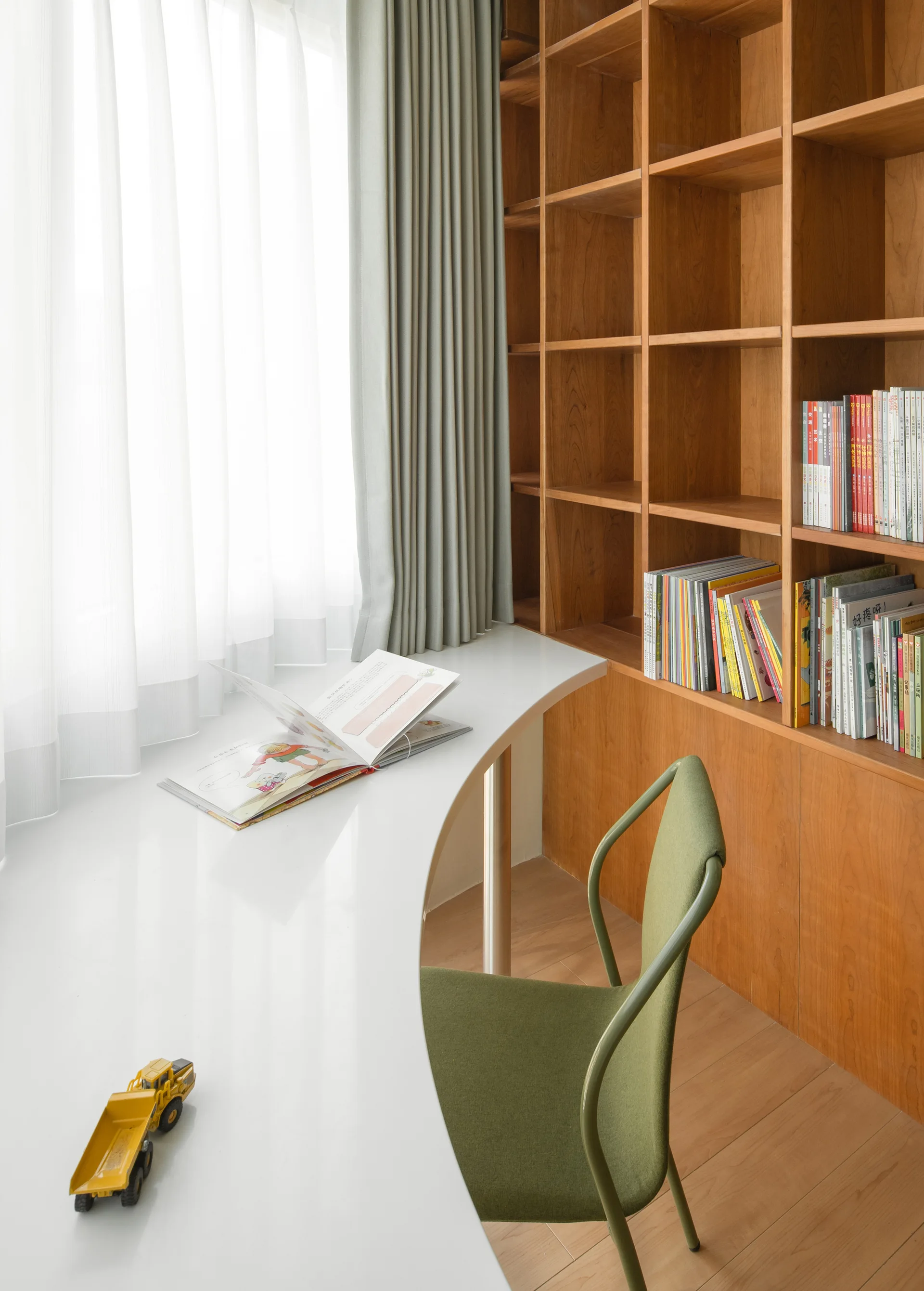 A bright study nook featuring a curved white desk, a sage green upholstered chair, and built-in floor-to-ceiling wooden bookshelves.