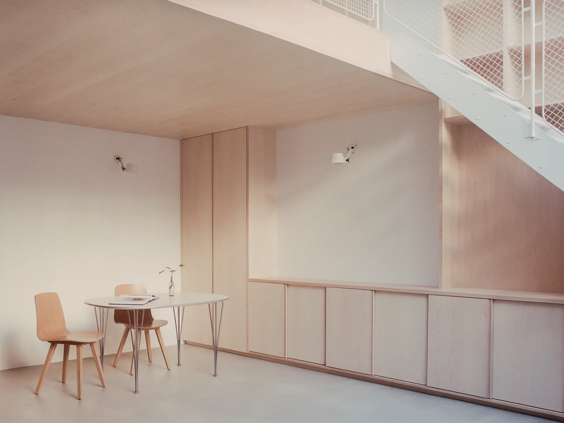 A minimalist dining area featuring a pale wood ceiling, built-in storage cabinets, a white oval table, and a staircase with white mesh railing.