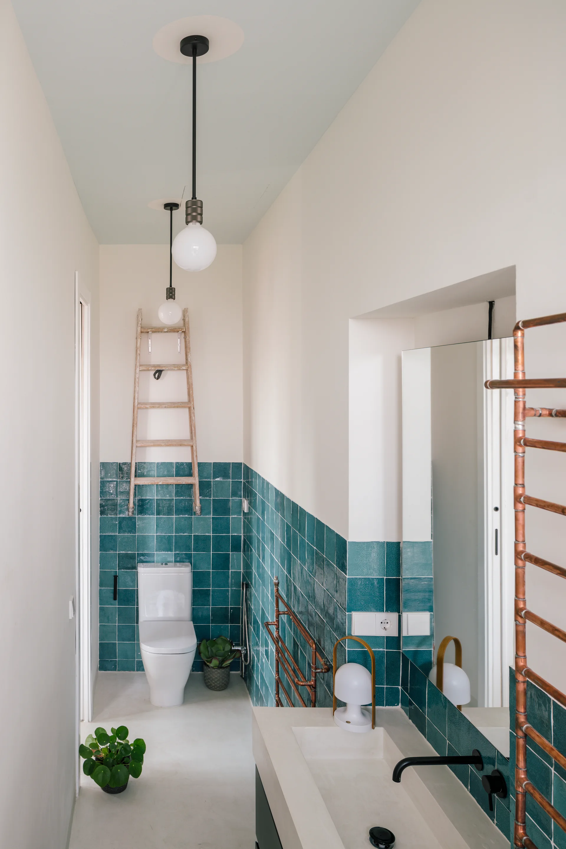 A narrow bathroom with teal square wall tiles, a pale blue ceiling, a concrete vanity, and a wooden ladder.