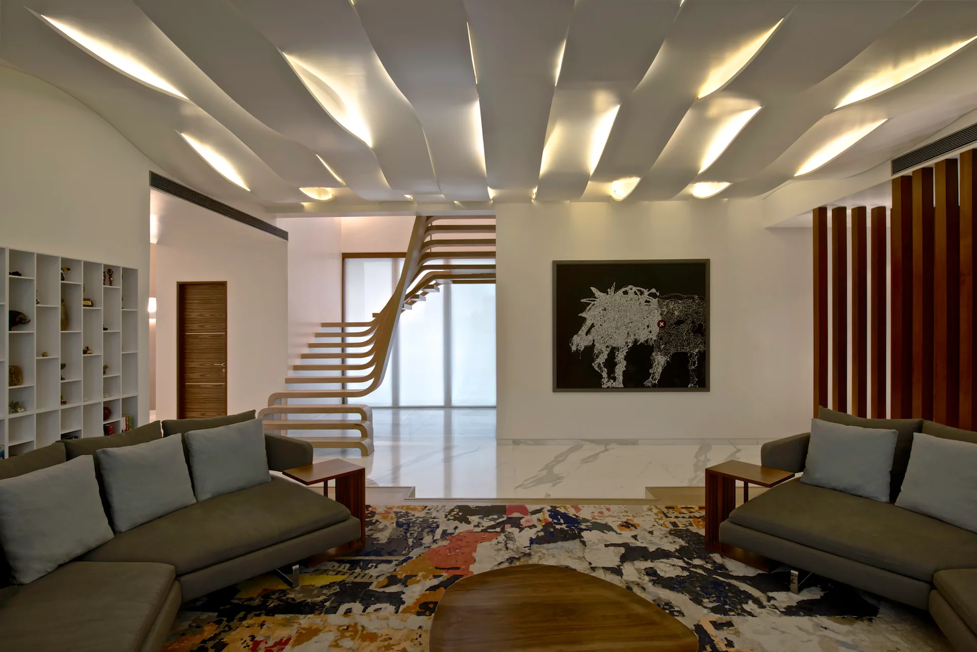 A modern living room features grey sofas on a patterned rug, a sculptural wavy ceiling with integrated lighting, and a floating wooden staircase.