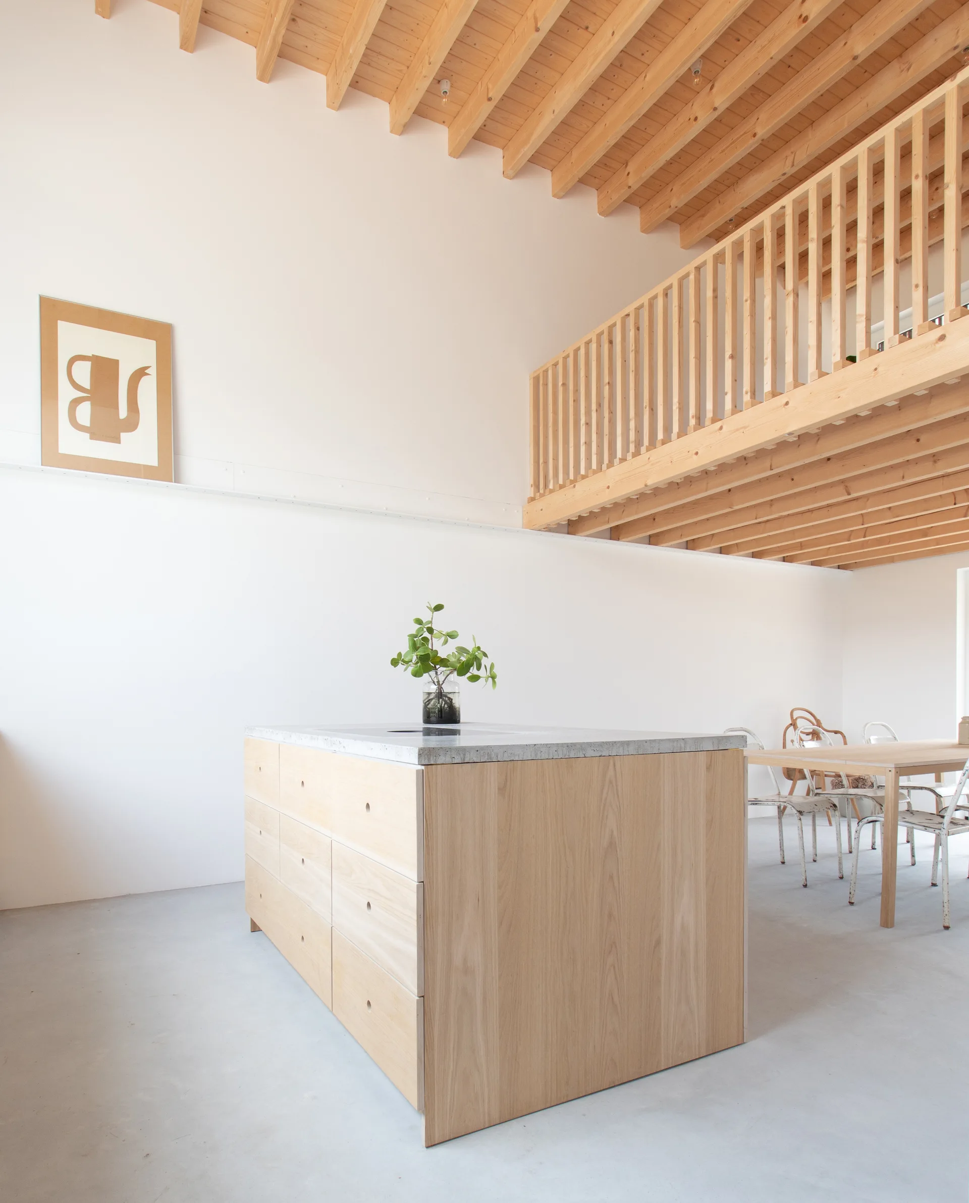 A minimalist kitchen features a light wood island with a concrete countertop, situated beneath a wooden loft mezzanine and exposed wood ceiling rafters.