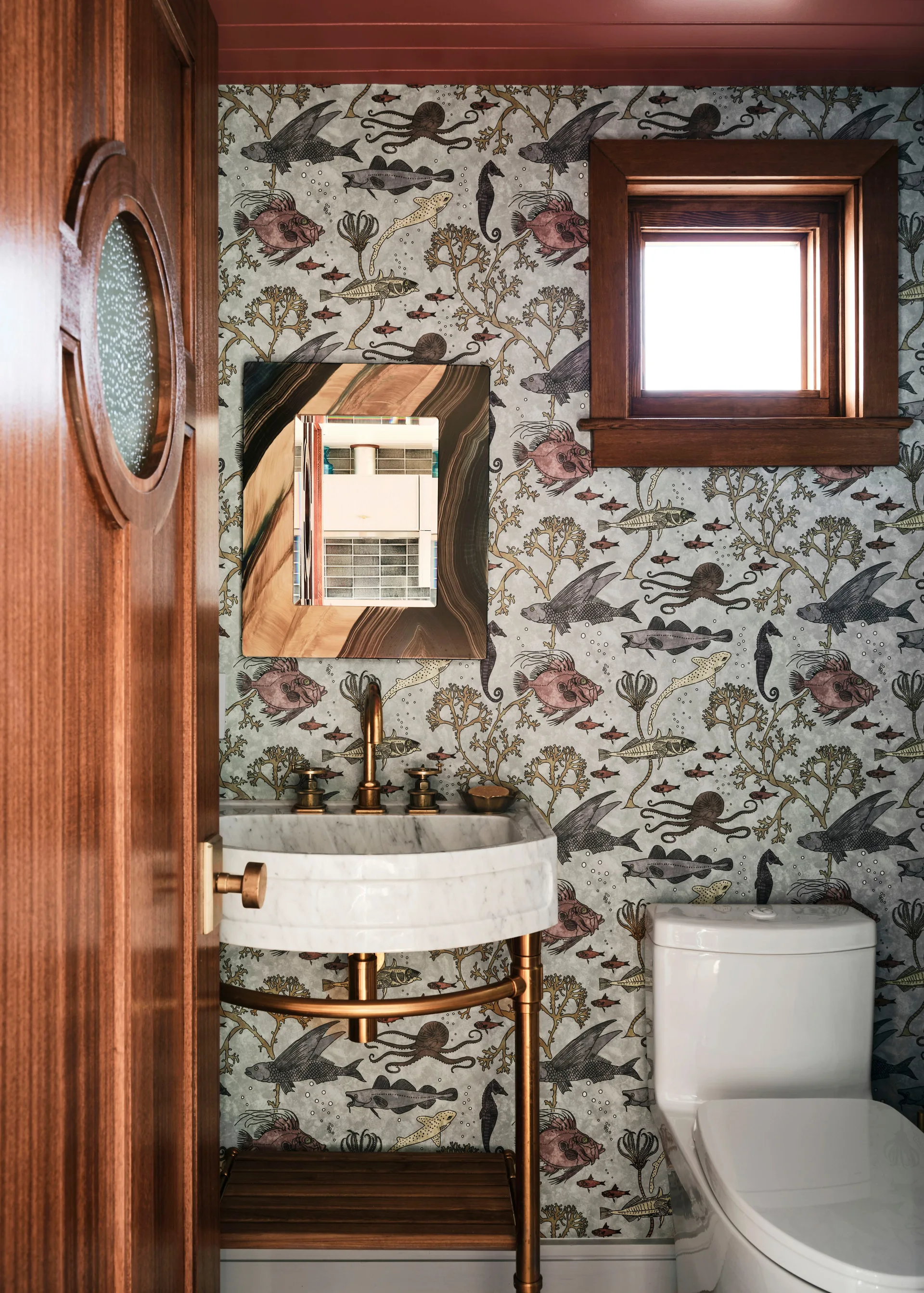 A powder room features marine-life patterned wallpaper, a marble console sink with brass legs, a wood door with a porthole, and a white toilet.