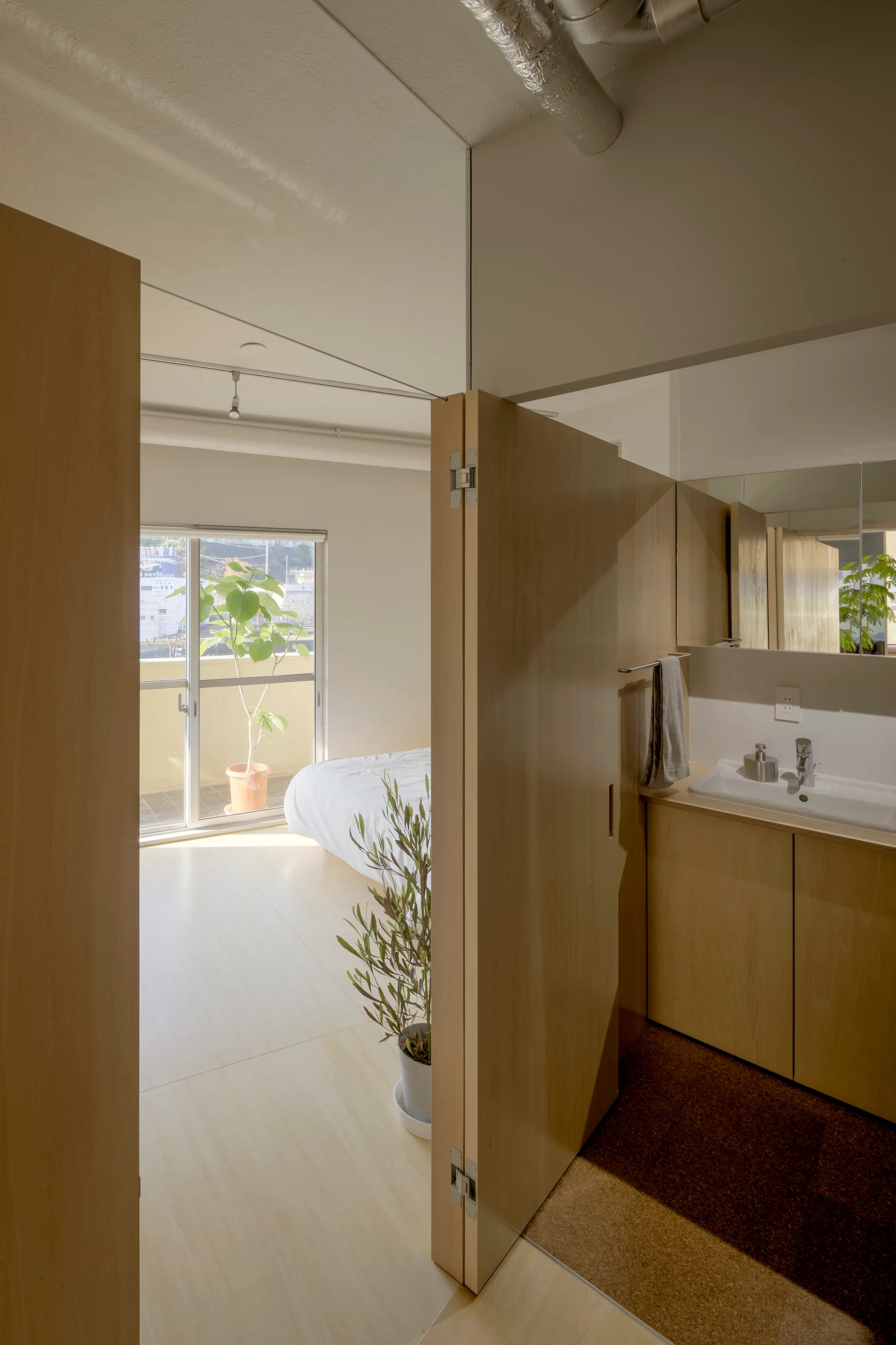 A folding wood partition door separates a bedroom with a large window from a bathroom vanity with a white sink and mirror.