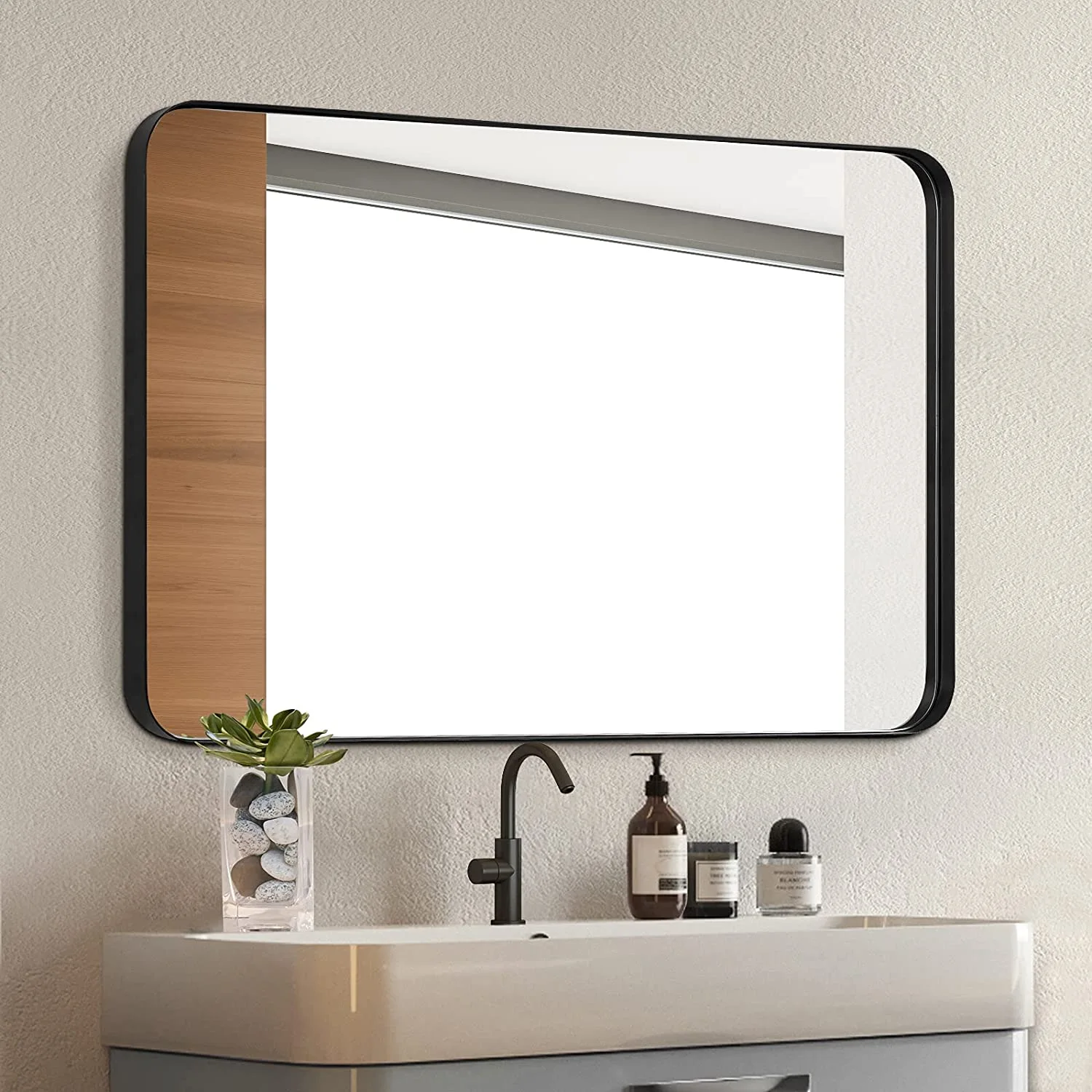 A rectangular wall mirror with a black metal frame and a vertical wood accent panel hangs above a light grey vanity and matte black faucet.