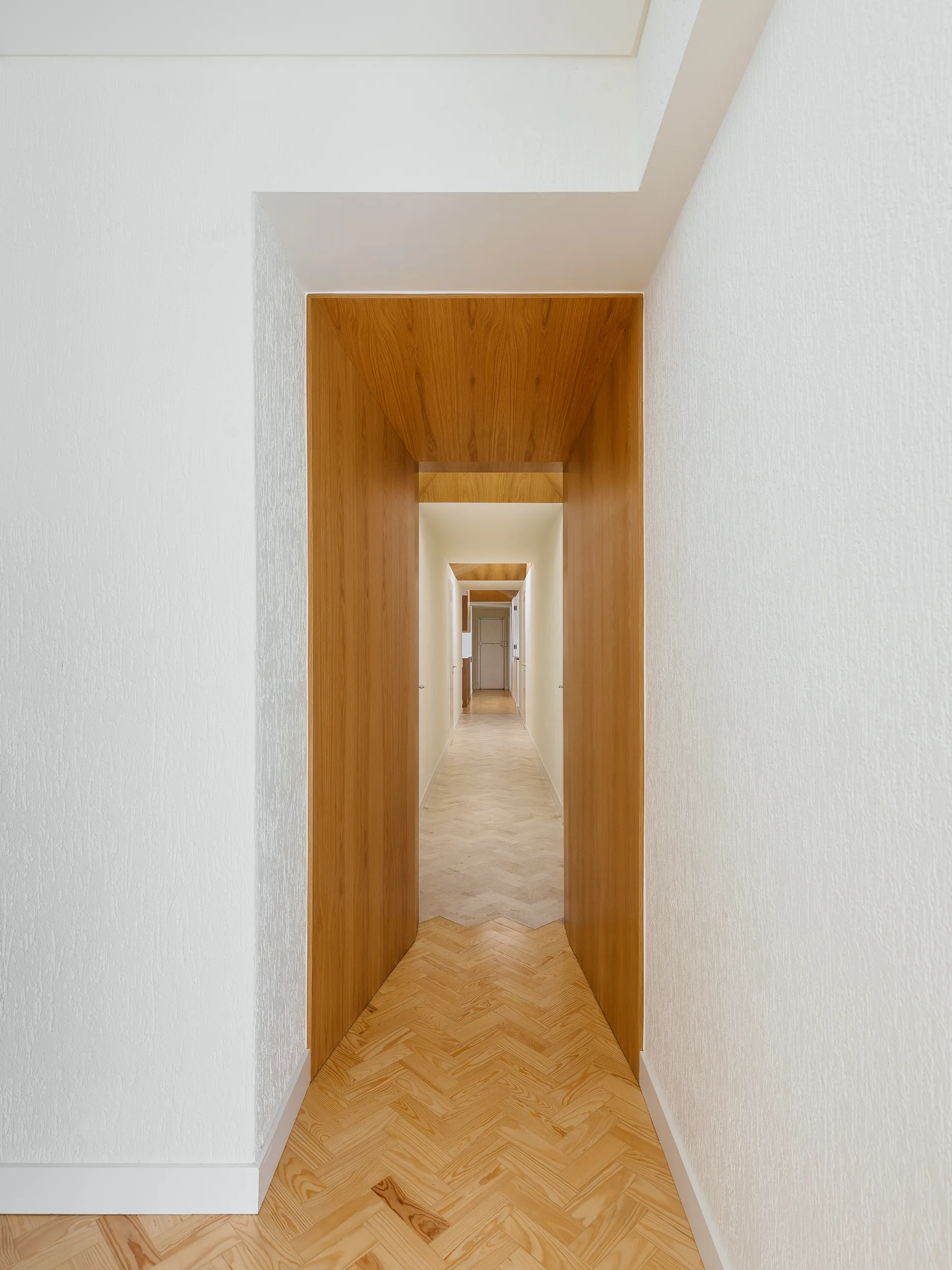 A long white hallway features light wood herringbone floors and a series of deep wood-lined portal openings leading into the distance.