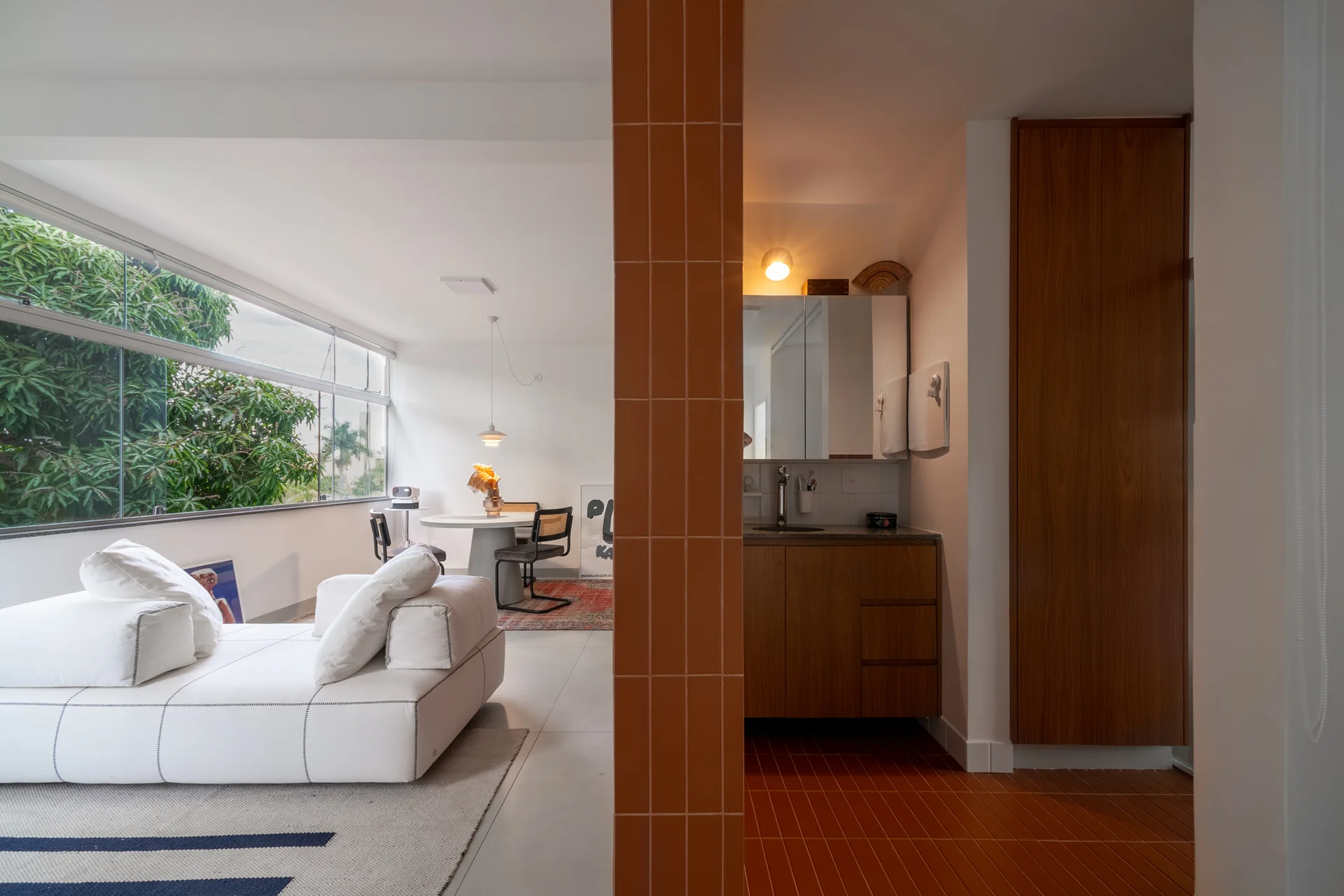 A modern open-plan living room features a white sofa, a dining table, and a terracotta-tiled partition next to a wooden vanity.