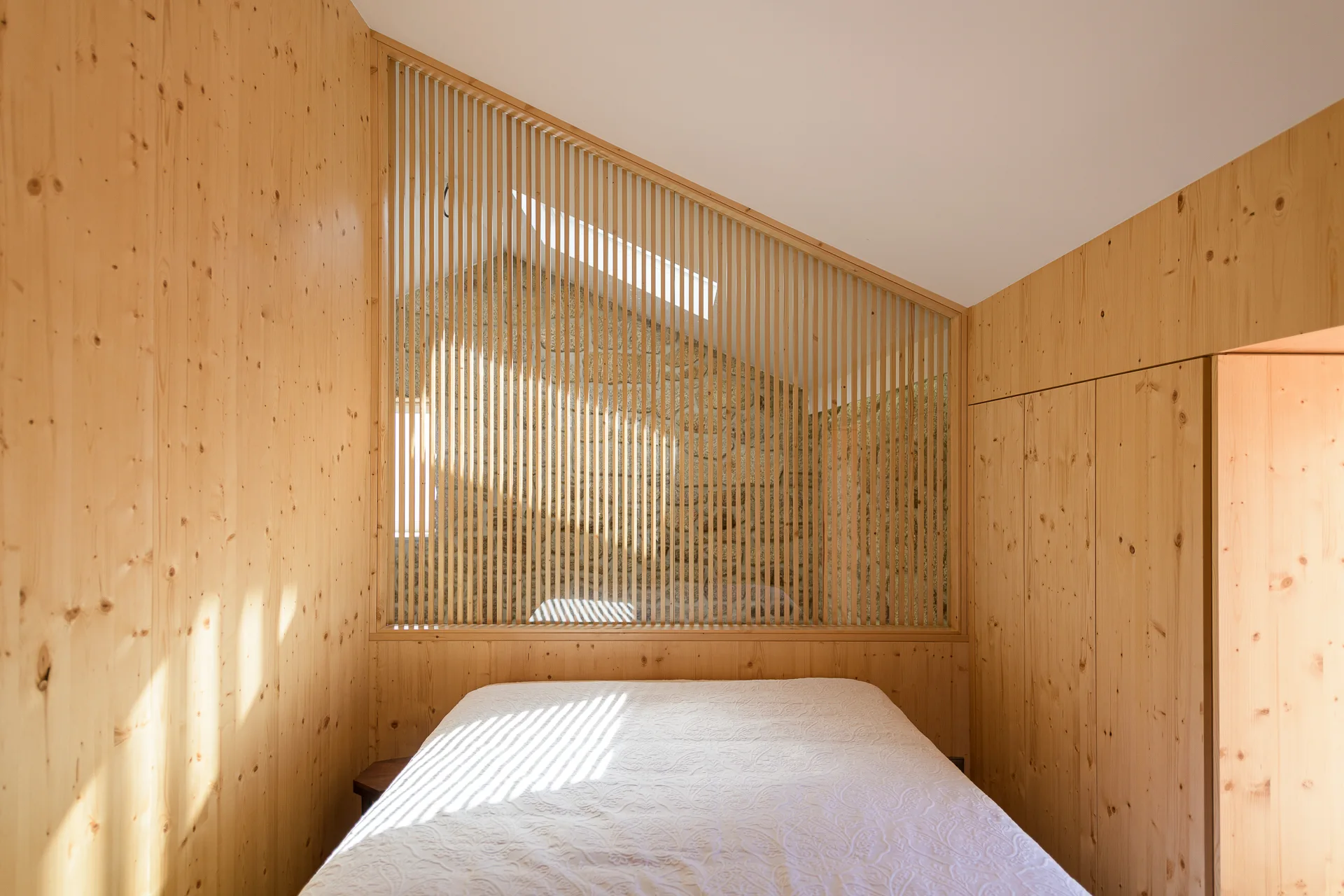 A minimalist bedroom featuring warm wood wall paneling, built-in closets, and a slatted timber partition screen in front of a stone wall.