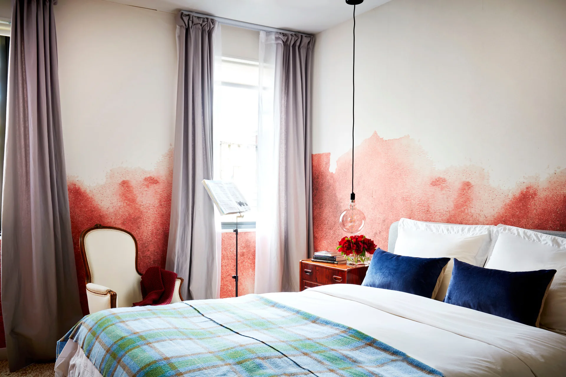 A bedroom features a bed with navy velvet pillows, a watercolor-effect terracotta mural, a vintage armchair, and grey drapes.
