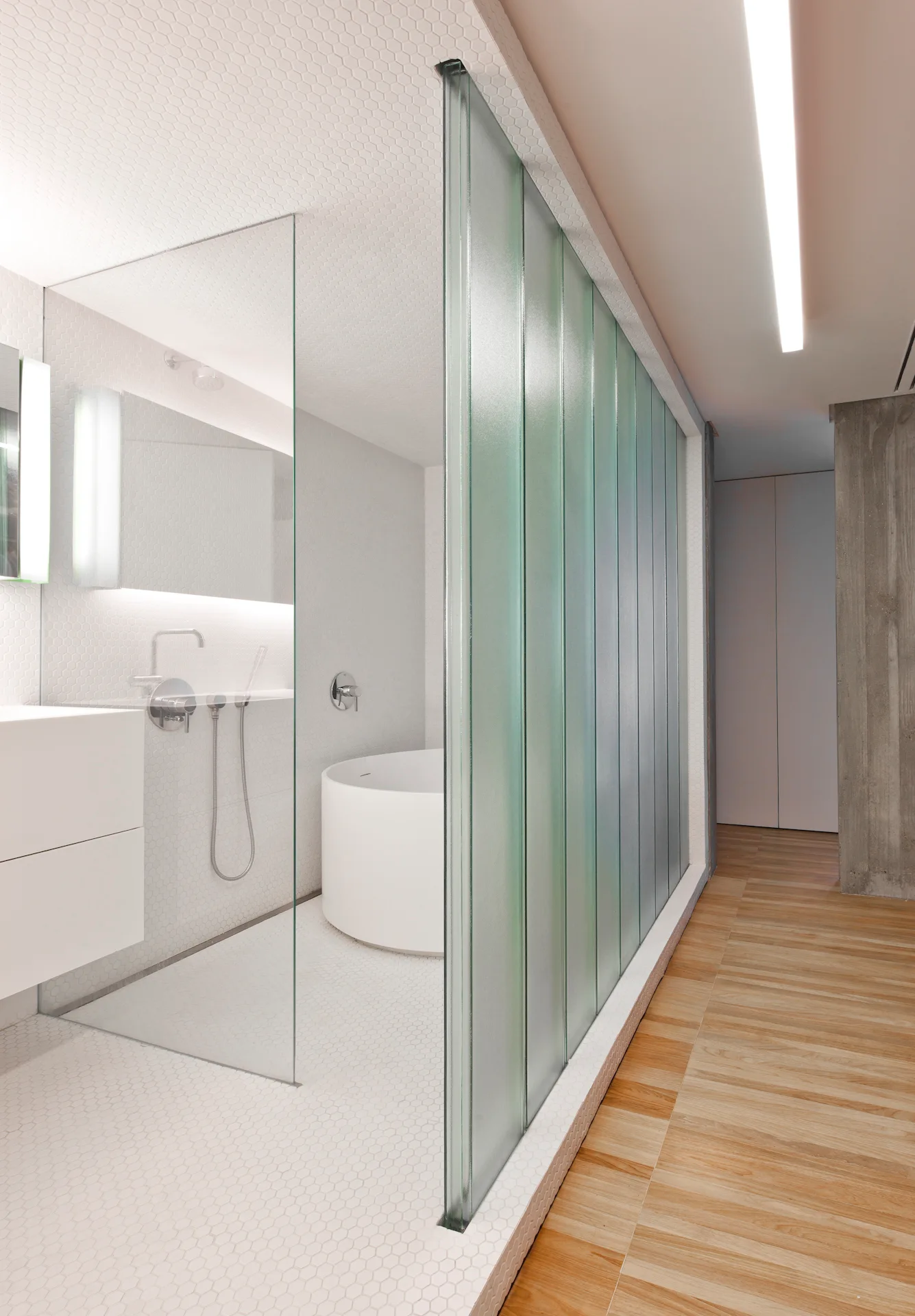 A modern bathroom featuring a white circular soaking tub, white hexagonal wall and floor tiles, and a textured channel glass partition.