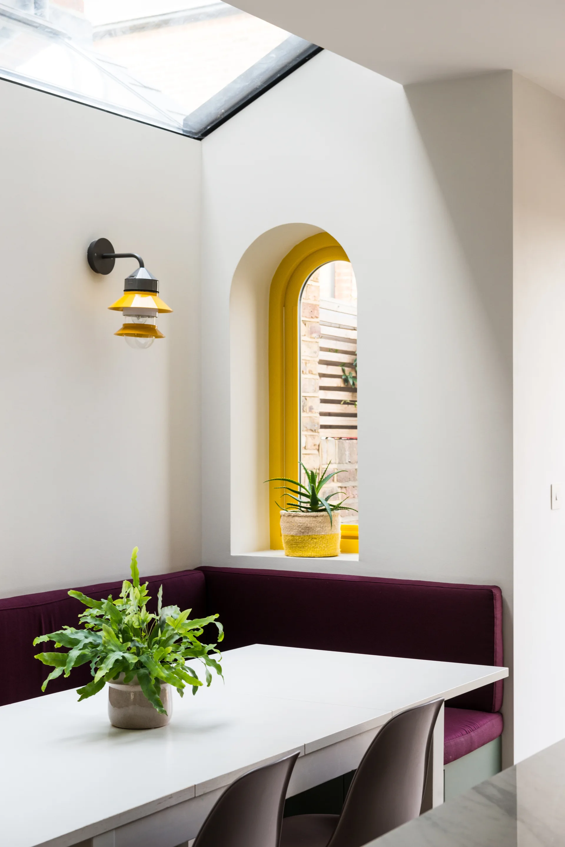 A dining nook features a plum-colored corner banquette under a skylight next to a white table and an arched window with a bright yellow frame.